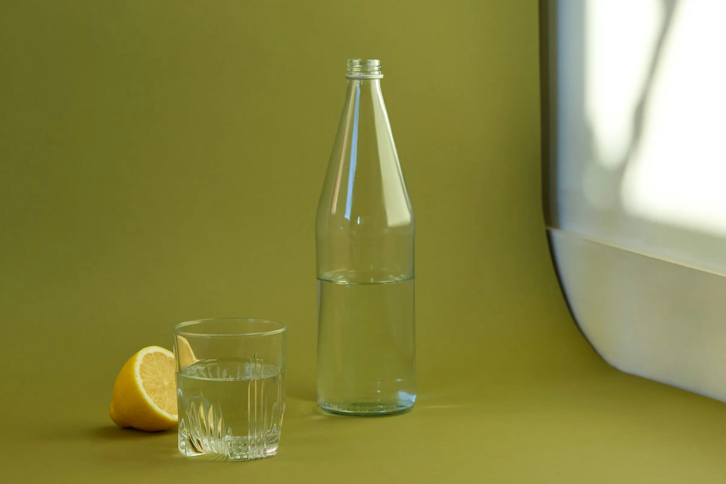 Glass bottle and tumbler of water with a lemon half on an olive green background.