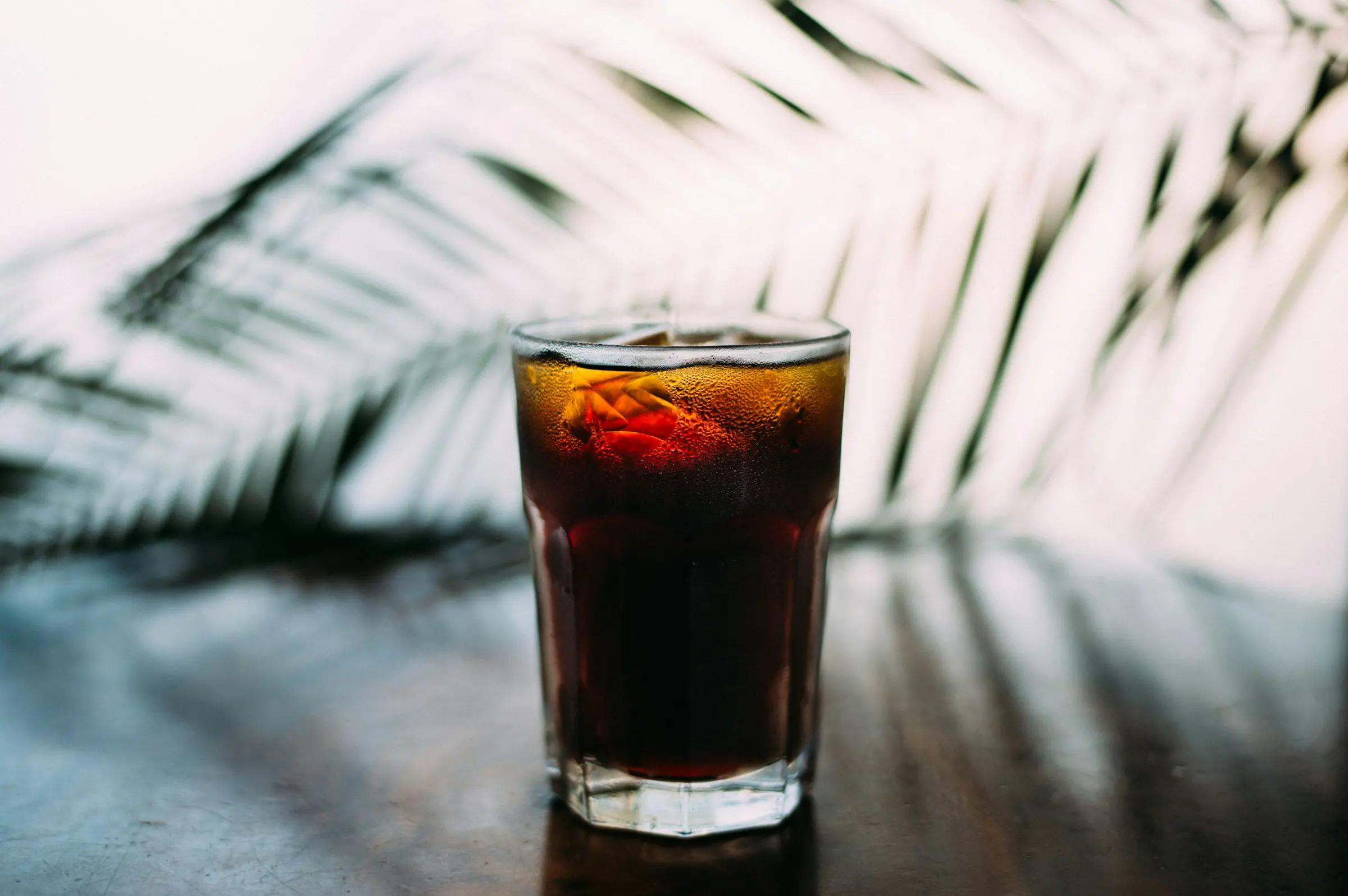 Glass of chilled soda on a wooden table with a blurred palm leaf backdrop.