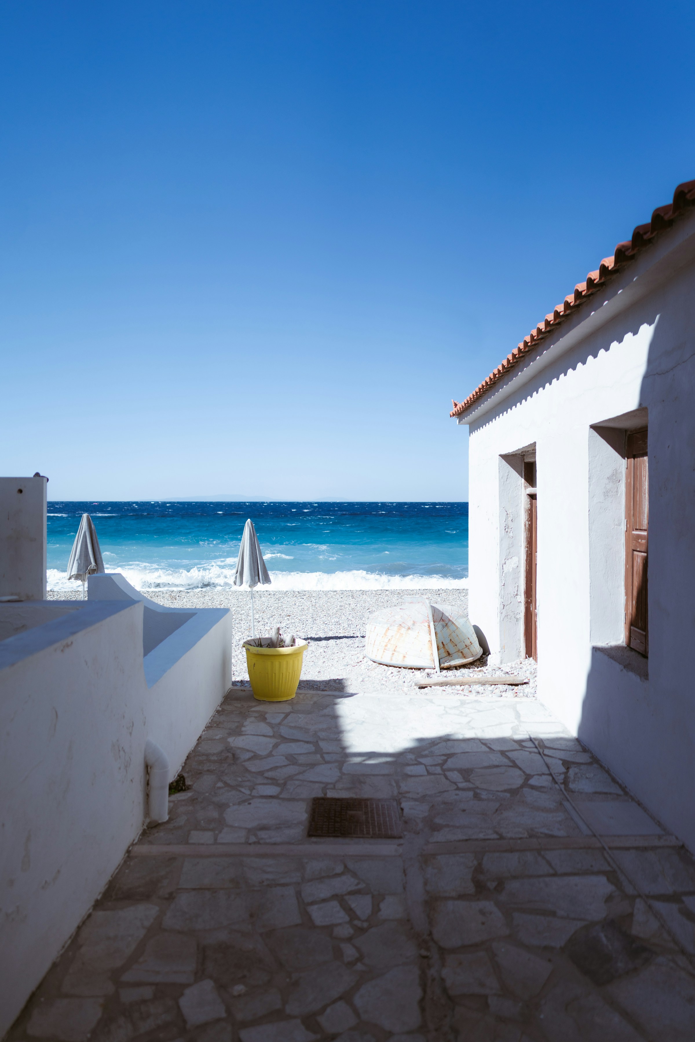 Vit medelhavsstil husfasad med stengolv och en öppen utsikt över en klarblå himmel och havet med strandparasoller på Samos i Grekland