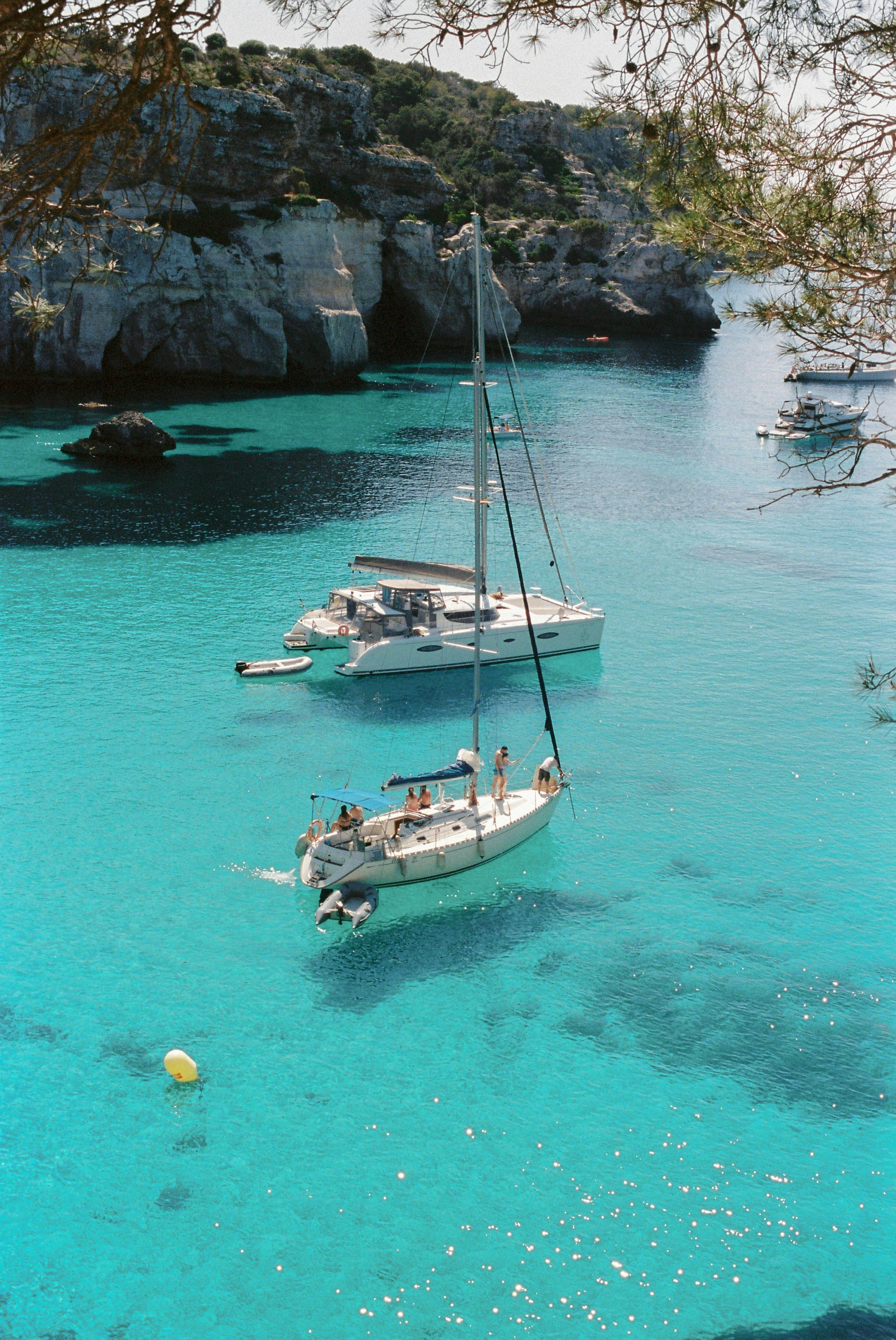 Segelbåtar förtöjda i en klar turkos vik omgiven av klippor på en solig dag på Menorca, Spanien.