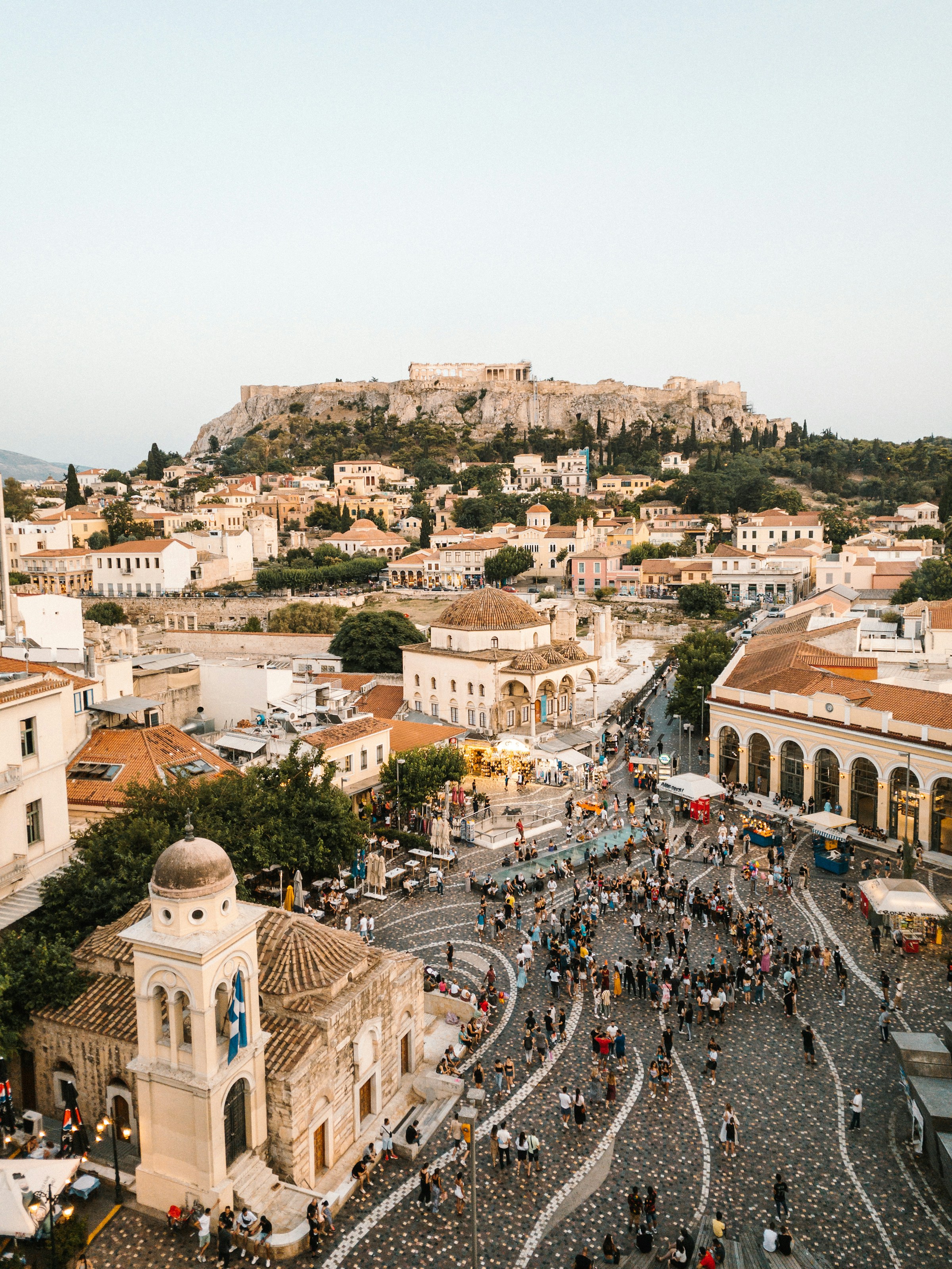 Flygfoto över ett myllrande torg med människor och klassisk arkitektur med en antik fästning i bakgrunden i Aten