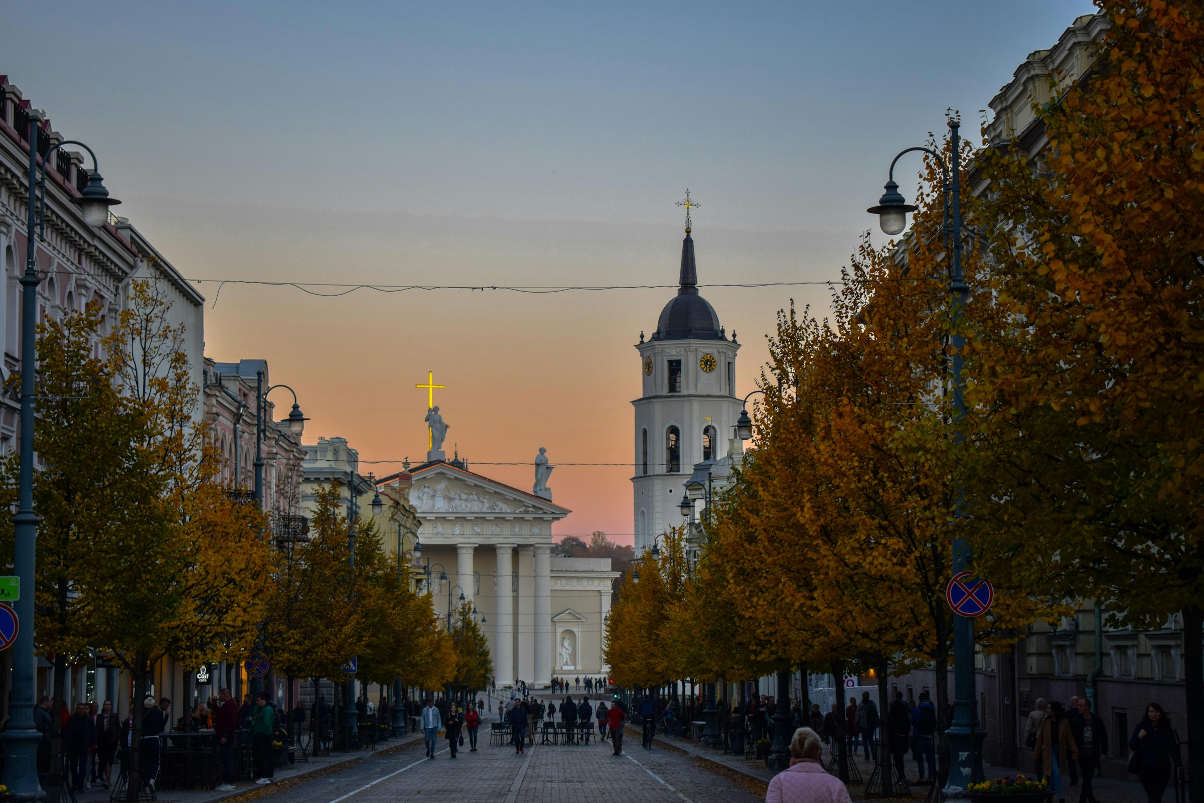 Stadsgata i skymningen med promenerande människor och höstfärgade lövträd, med en klassisk vit kyrka i bakgrunden i Vilnius.