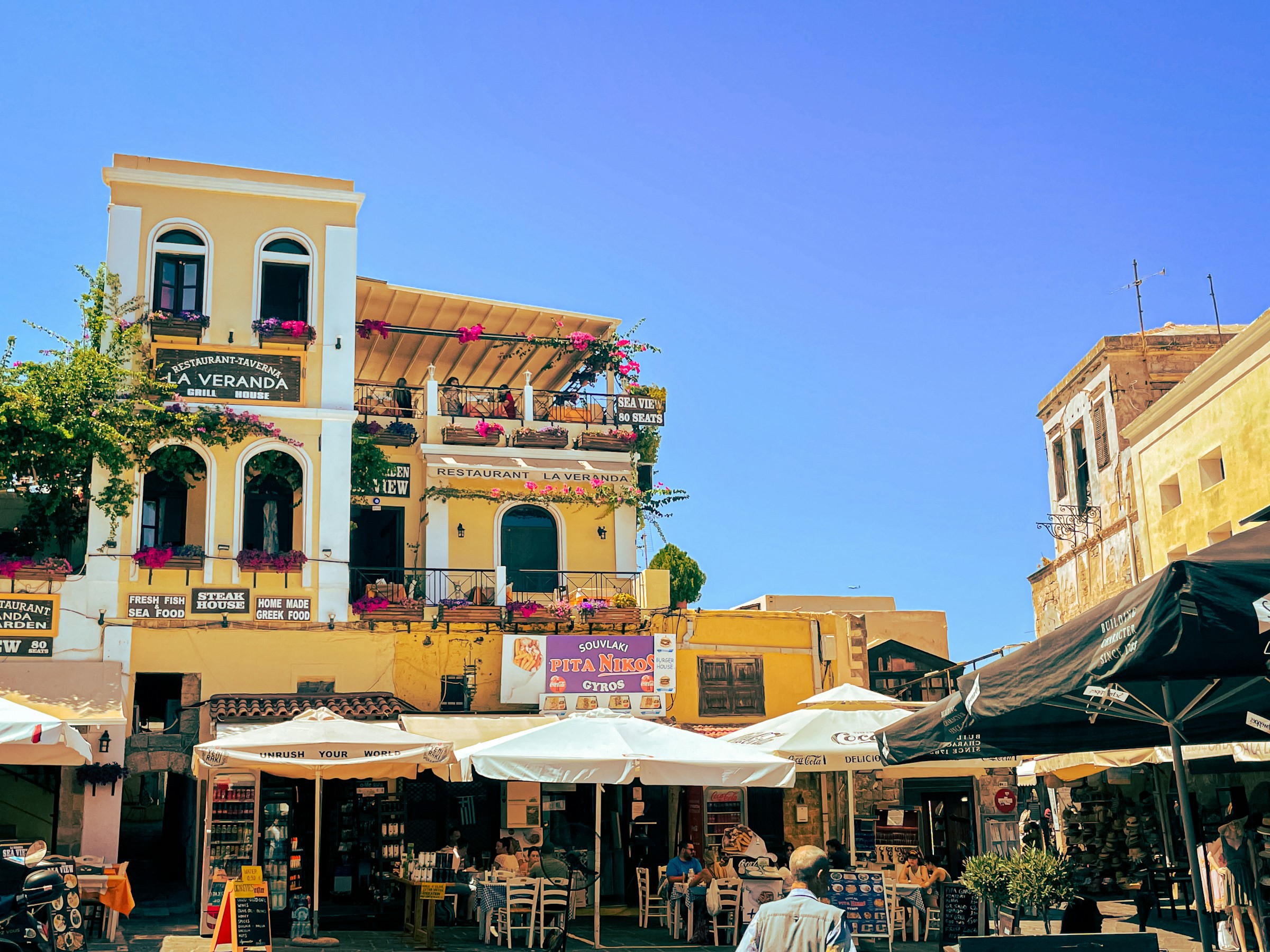 Livligt uteserveringsområde med restaurangterrass och blomsterdekorerade balkonger under en klarblå himmel på Rhodos, Grekland