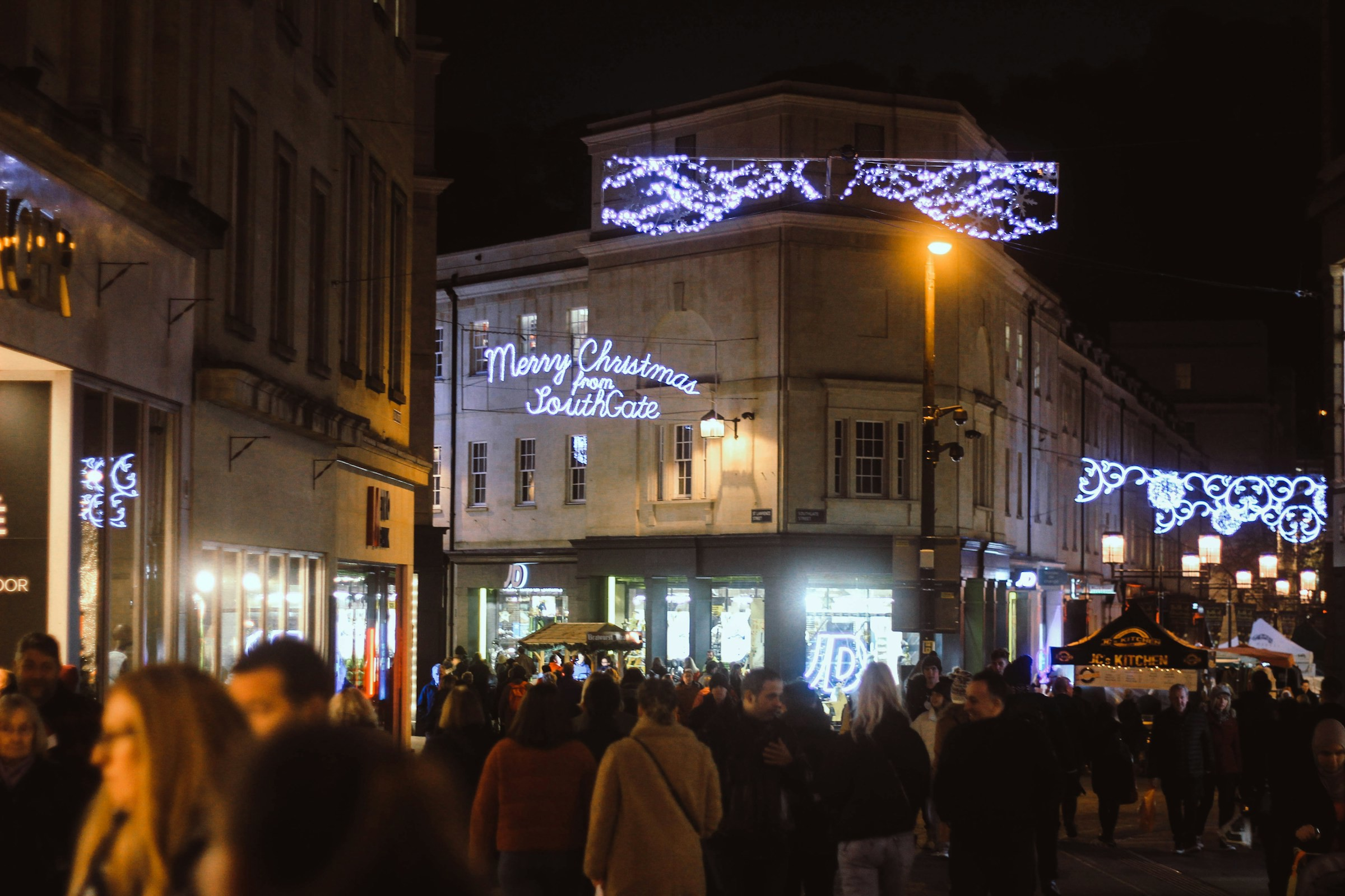 Julstämning på stadsgatan i Bath, Storbritannien med människor och upplysta skyltar som önskar 'God Jul från SouthGate'.