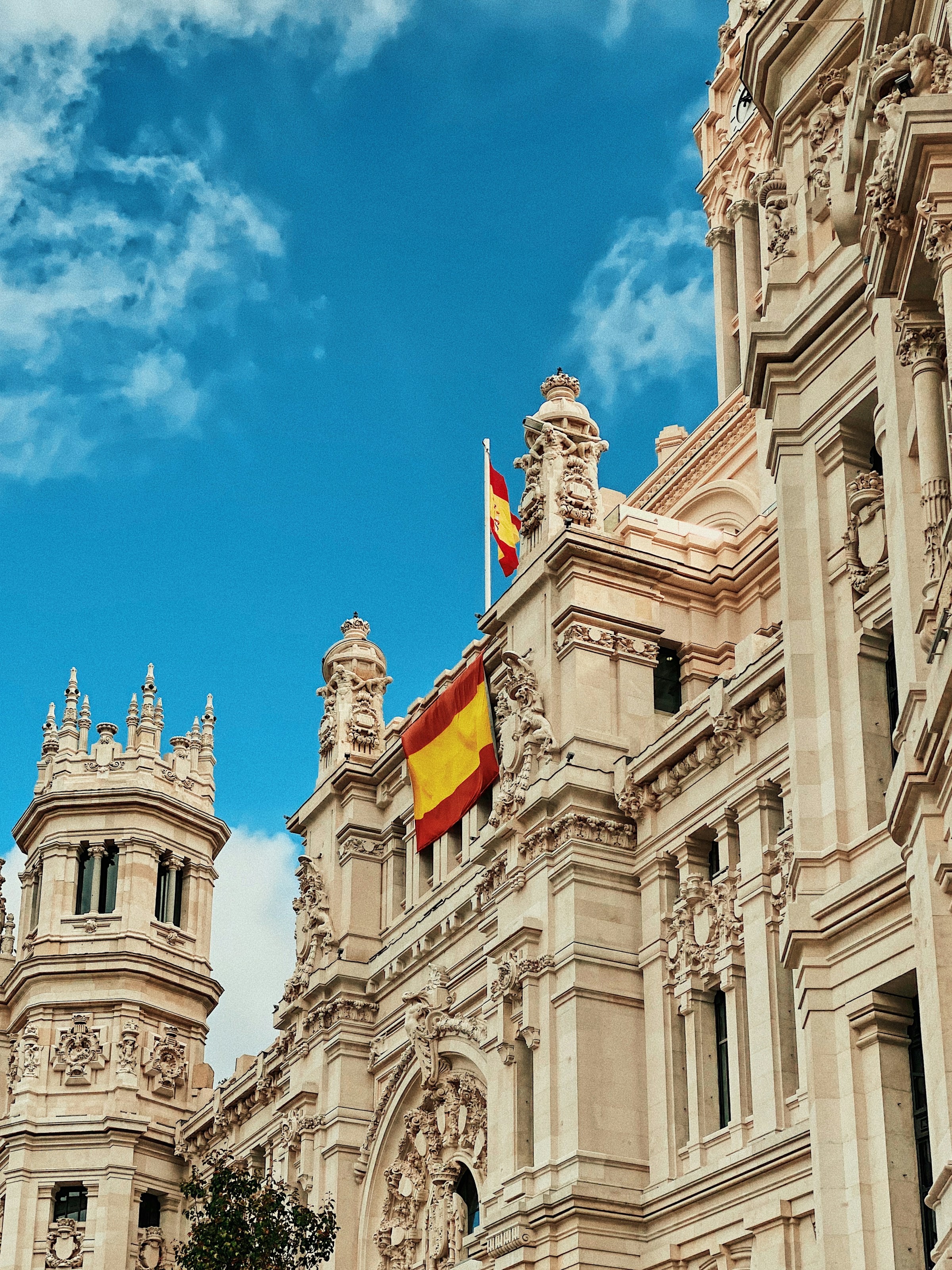 Spansk barockarkitektur och fladdrande spansk flagga mot en blå himmel med moln i Madrid.