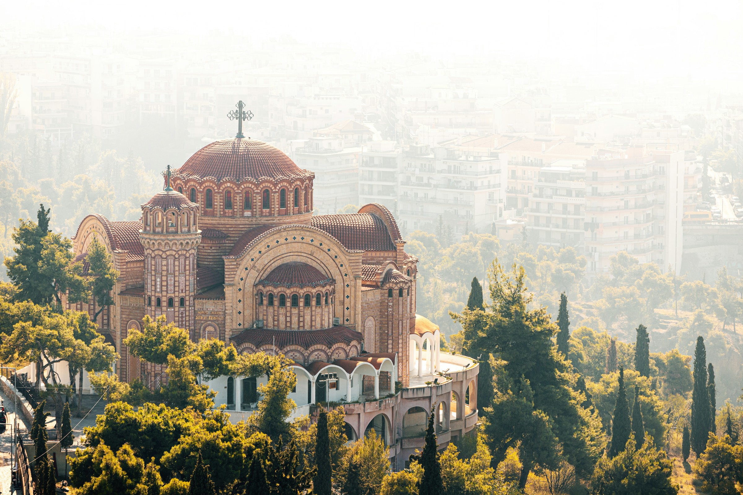 Kyrka med tegelarkitektur i en solbelyst stad med träd och byggnader i bakgrunden, beläget i ett kuperat landskap i Thessaloniki