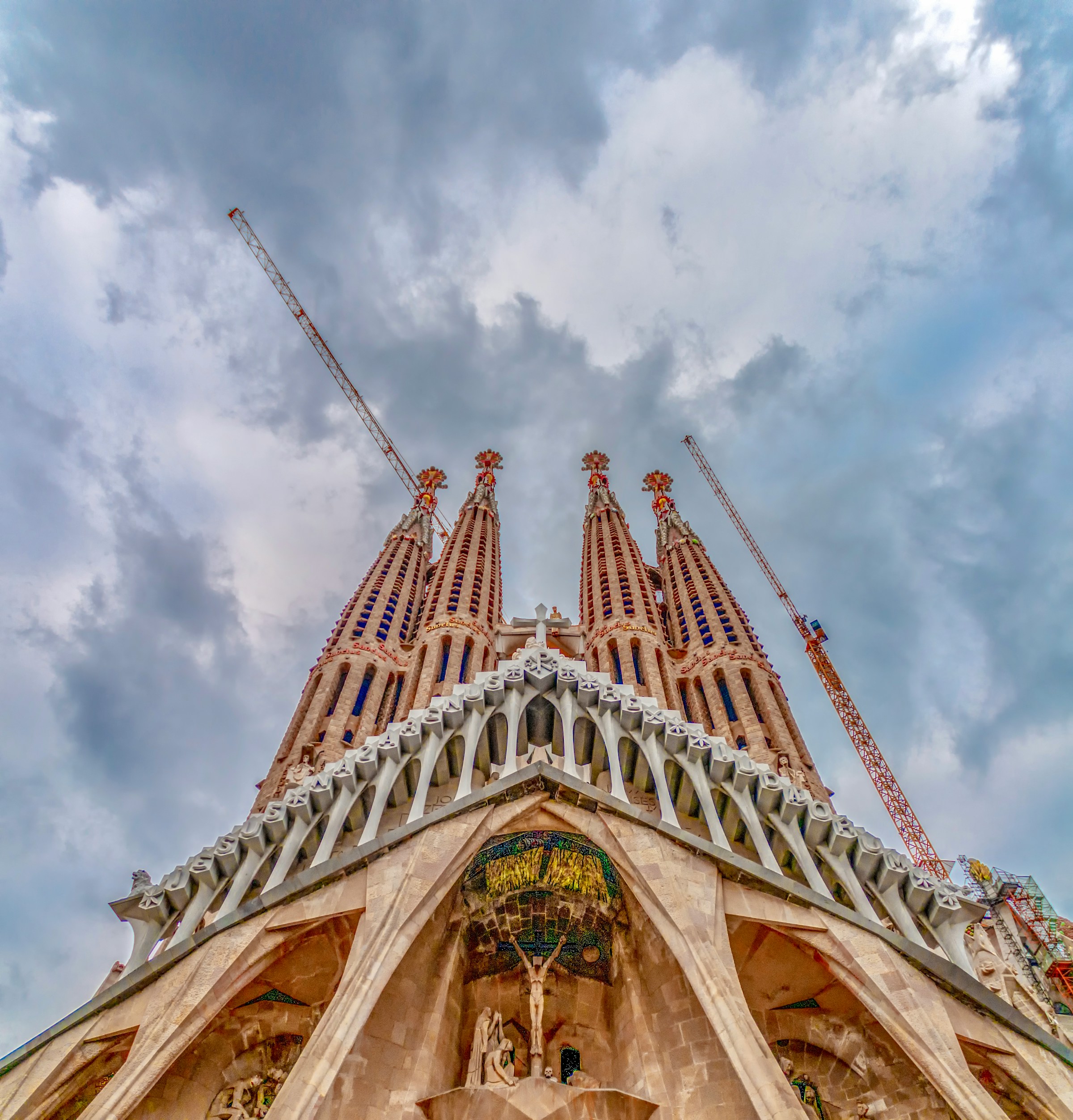 Dramatisk låg vinkel vy av Sagrada Familia i Barcelona med intrikata spiror och byggnadskranar mot en molnig himmel.