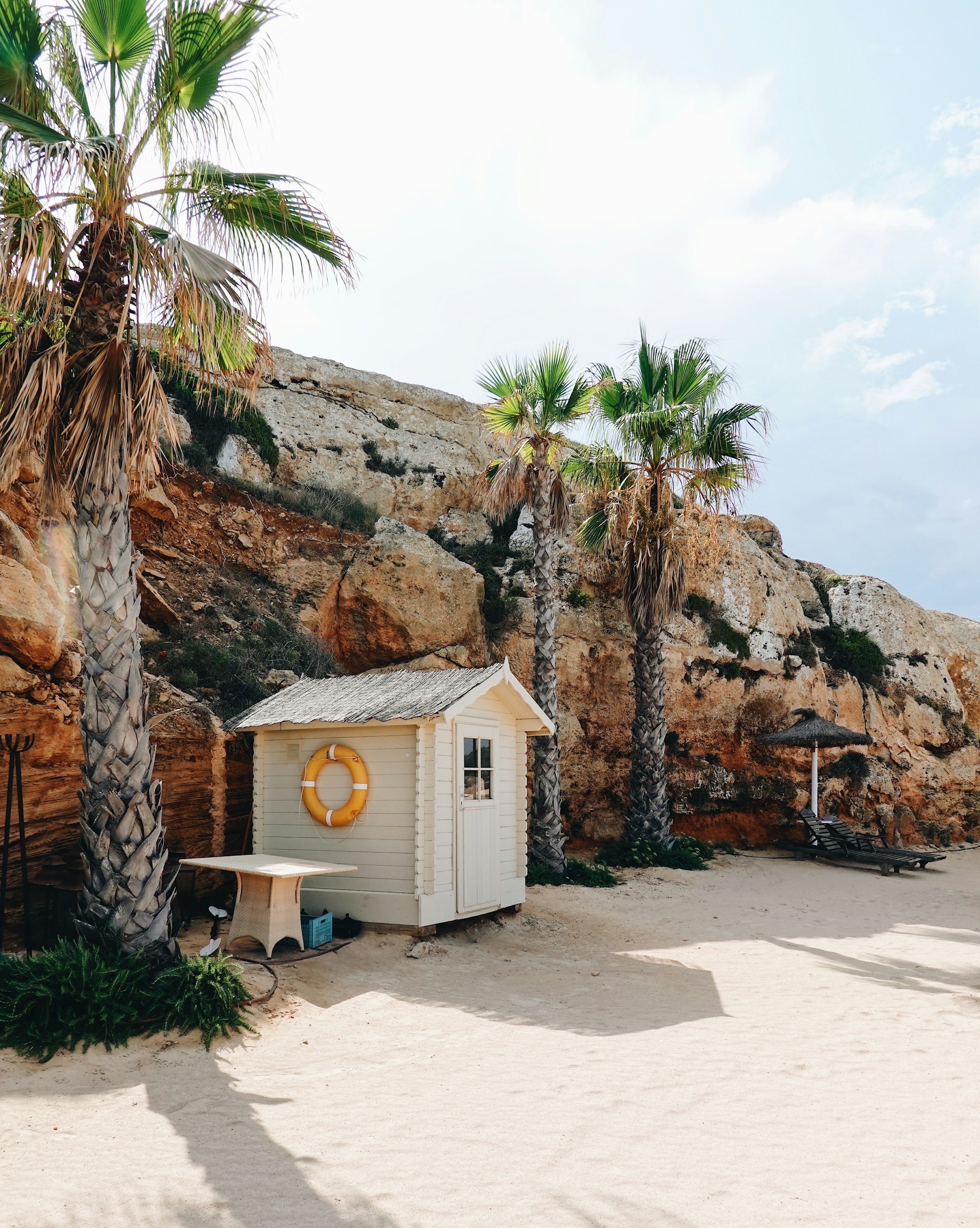 Vit strandstuga med livboj nära palmer och klippiga klippor på en solig dag på Mallorca.