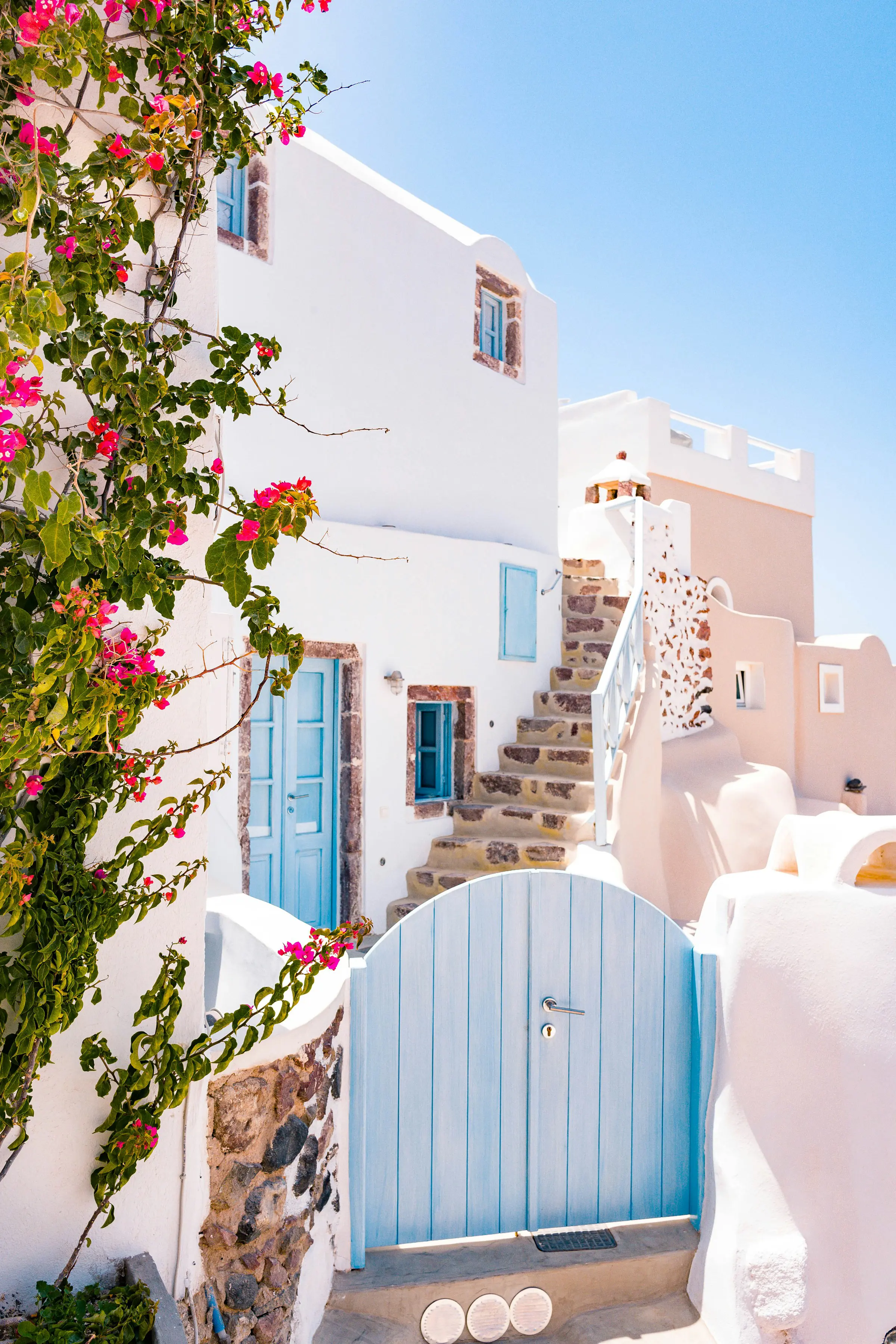 Whitewashed Greek house with blue door and gate, stone steps, and vibrant pink bougainvillea in summer.