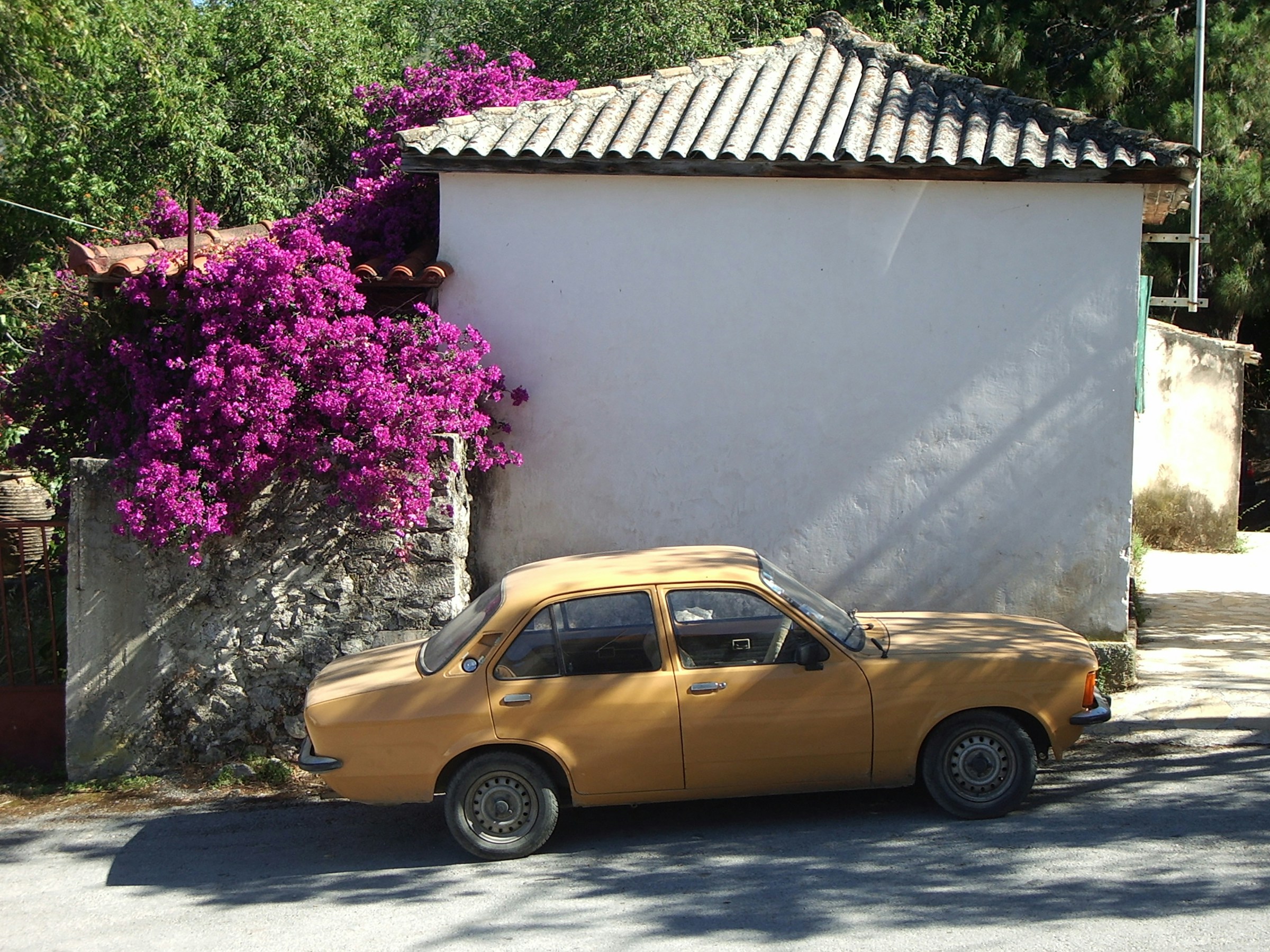 Gul veteranbil parkerad bredvid en vit vägg med lila bougainvillea i blom ovanför i soligt landskap på Zakynthos