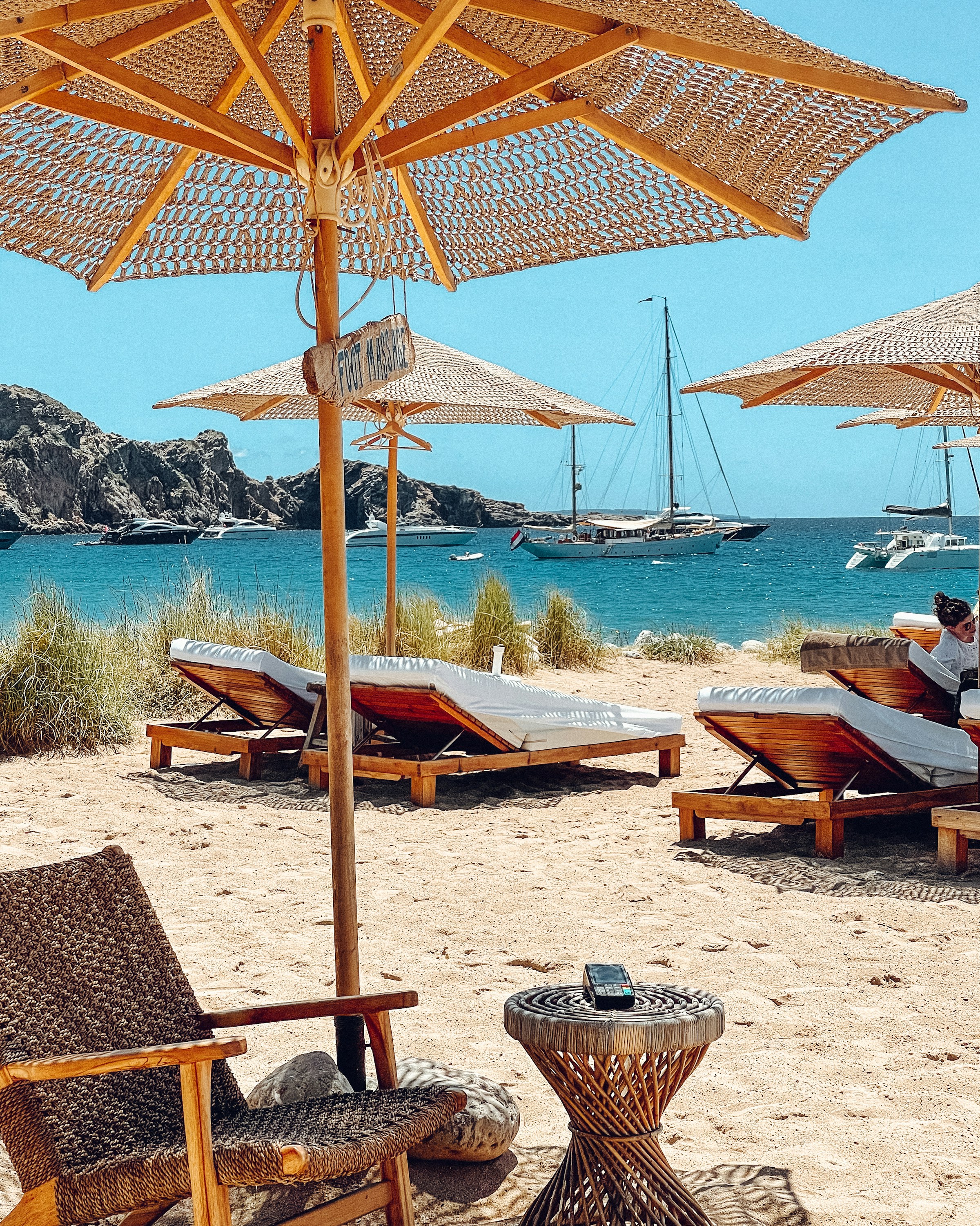 Strandparasoller och solstolar med utsikt över segelbåtar på klart vatten på Ibiza.
