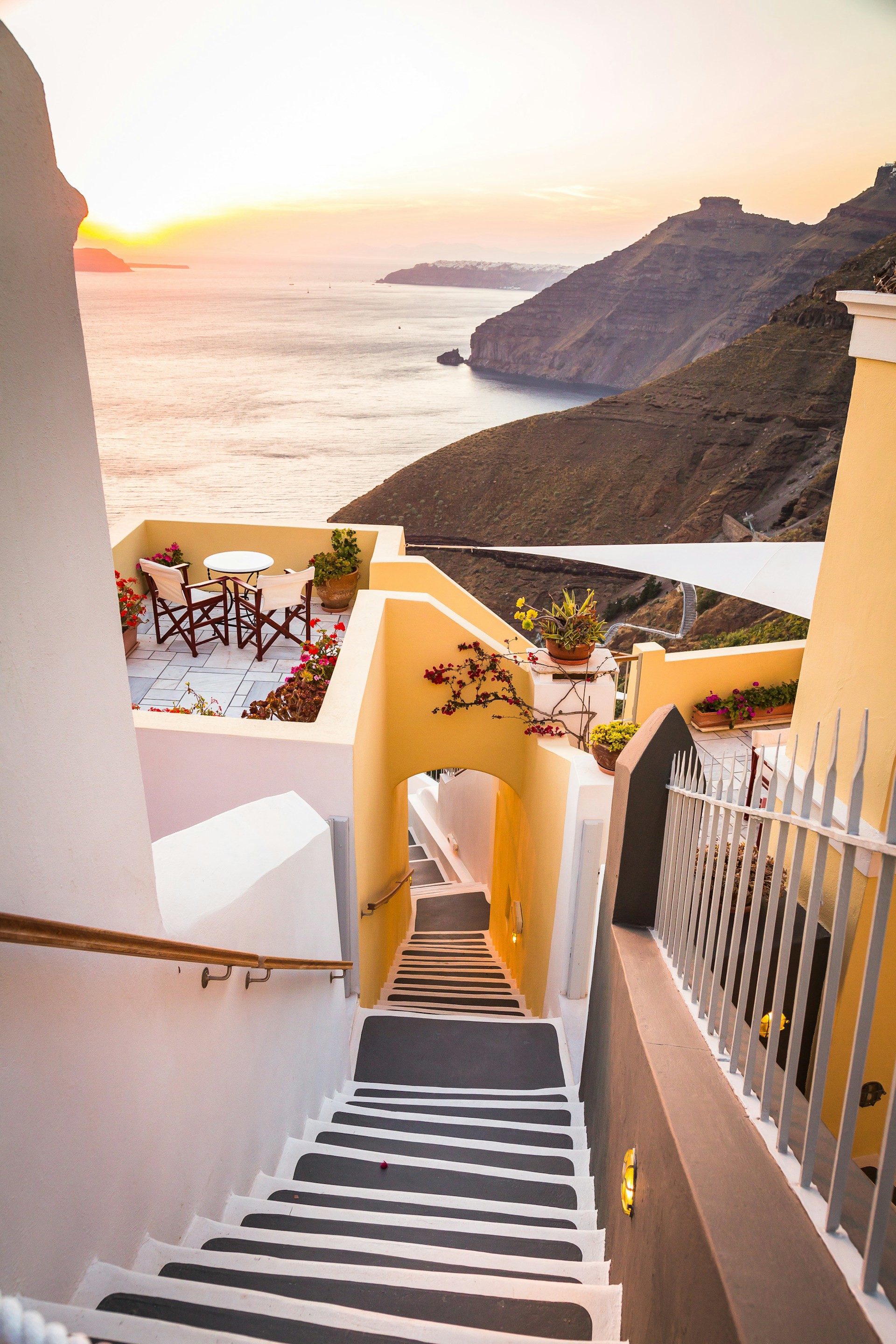 Vit och gul trappa som leder ned till en mysig uteservering med vy över Egeiska havet vid solnedgången, Santorini, Grekland.