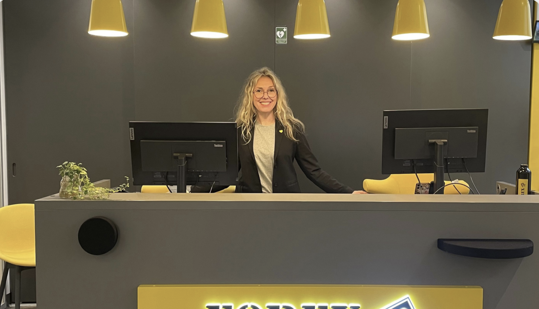 Leende receptionist vid incheckningsdisk med gul inredningsdetaljer och datorskärmar på ett modernt kontor.