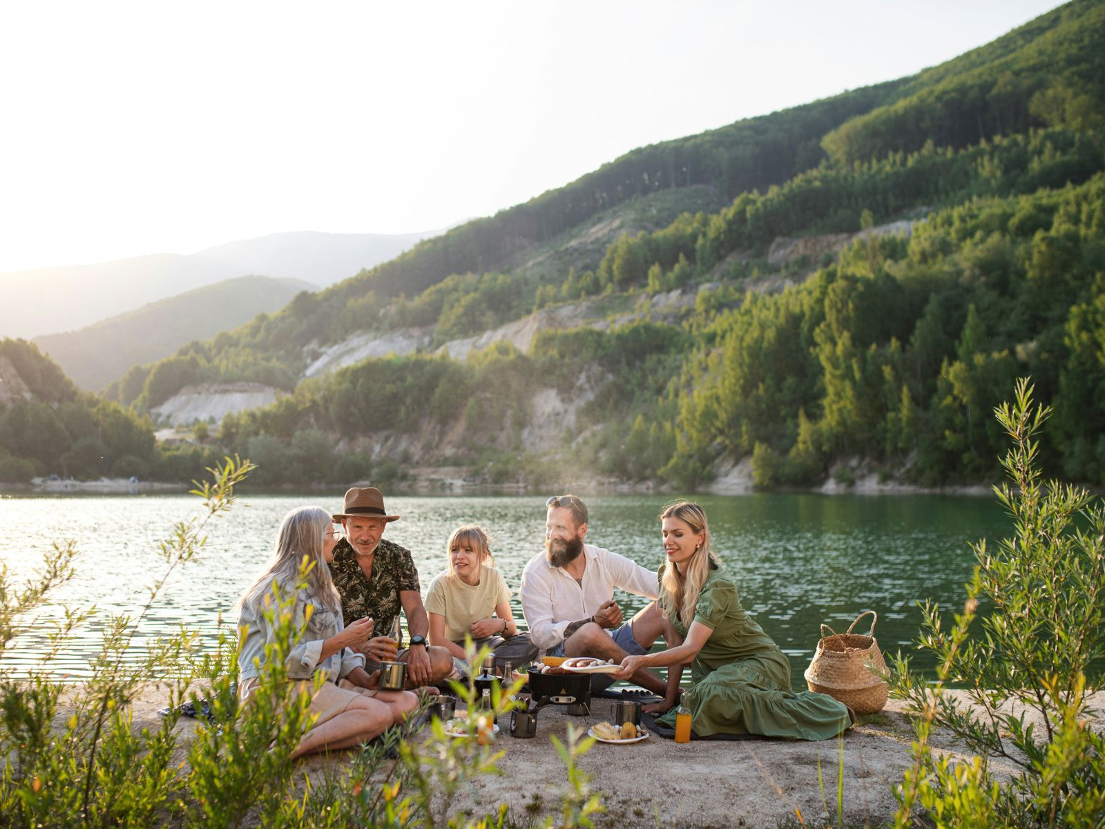 en grupp med vänner sitter vid en sjö och umgås med vackra berg i bakgrunden