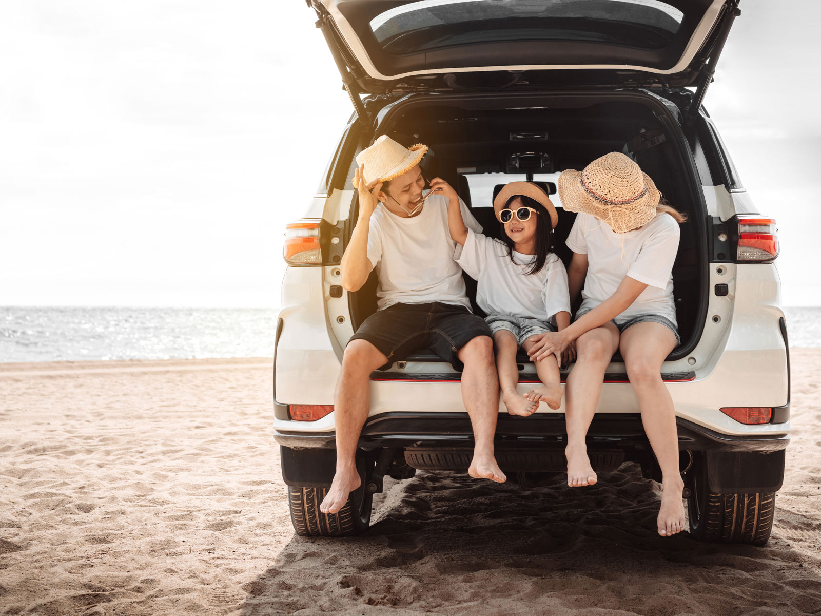 Jämför reseförsäkring - Tre personer som sitter i bagageutrymmet på en SUV vid stranden, bär solhattar och njuter av semestern.