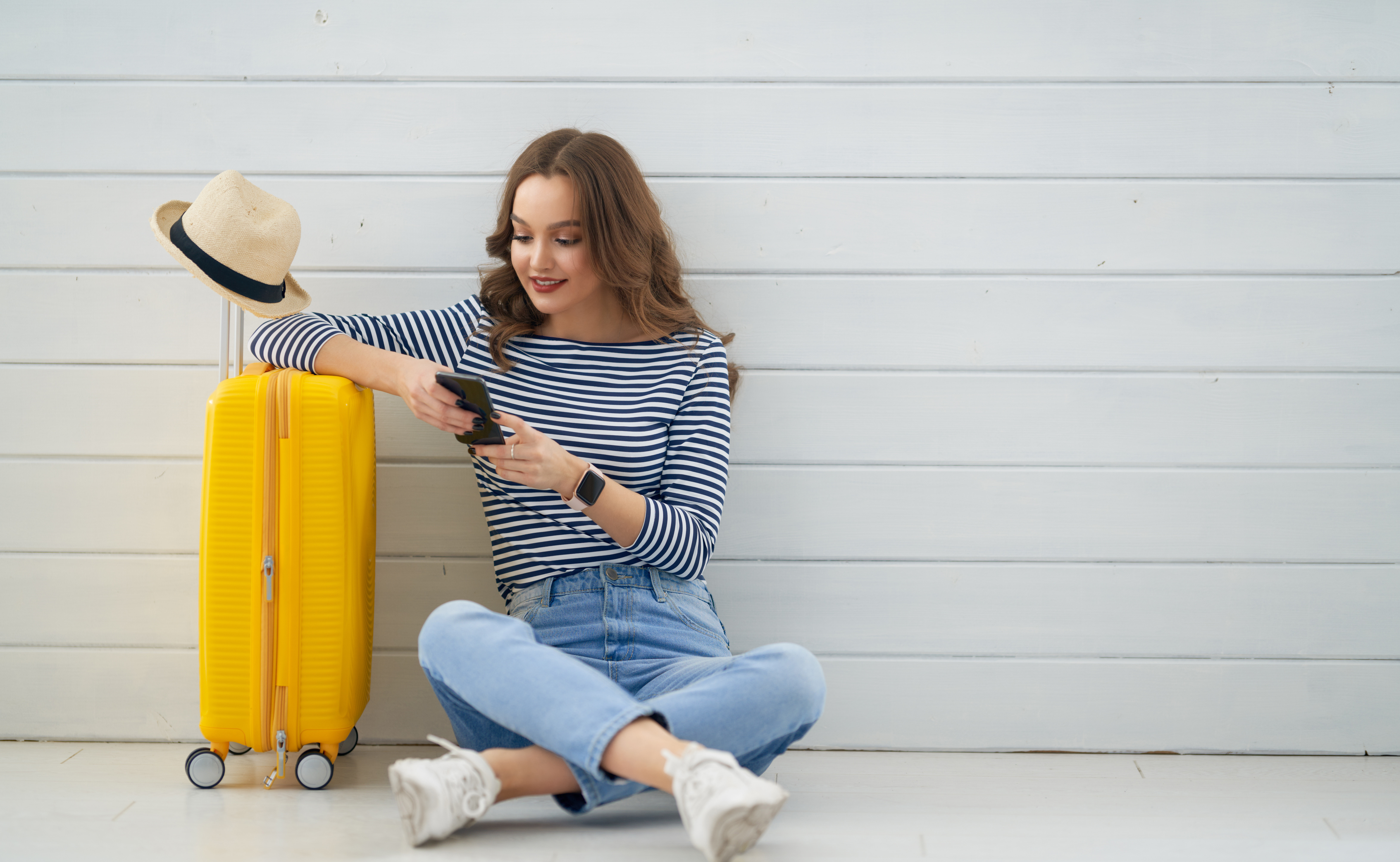 Bagageförsäkring - Kvinna med blårandig tröja och jeans sitter på golvet och använder sin smartphone bredvid en gul resväska.