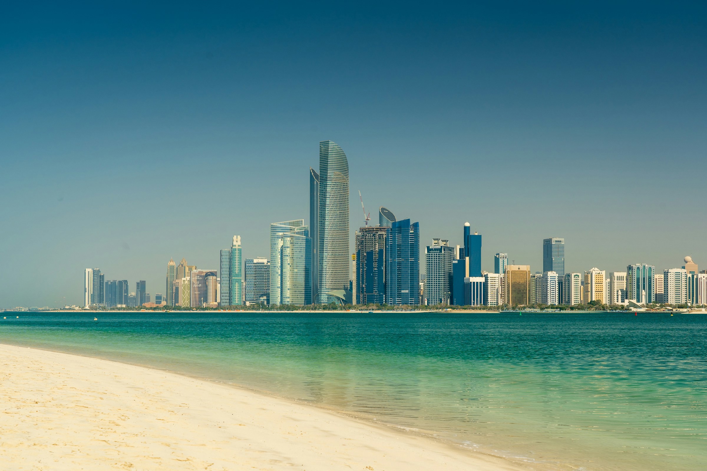 Vy över Abu Dhabi med höga skyskrapor från en vit sandstrand vid klart turkosblått hav under en klarblå himmel.