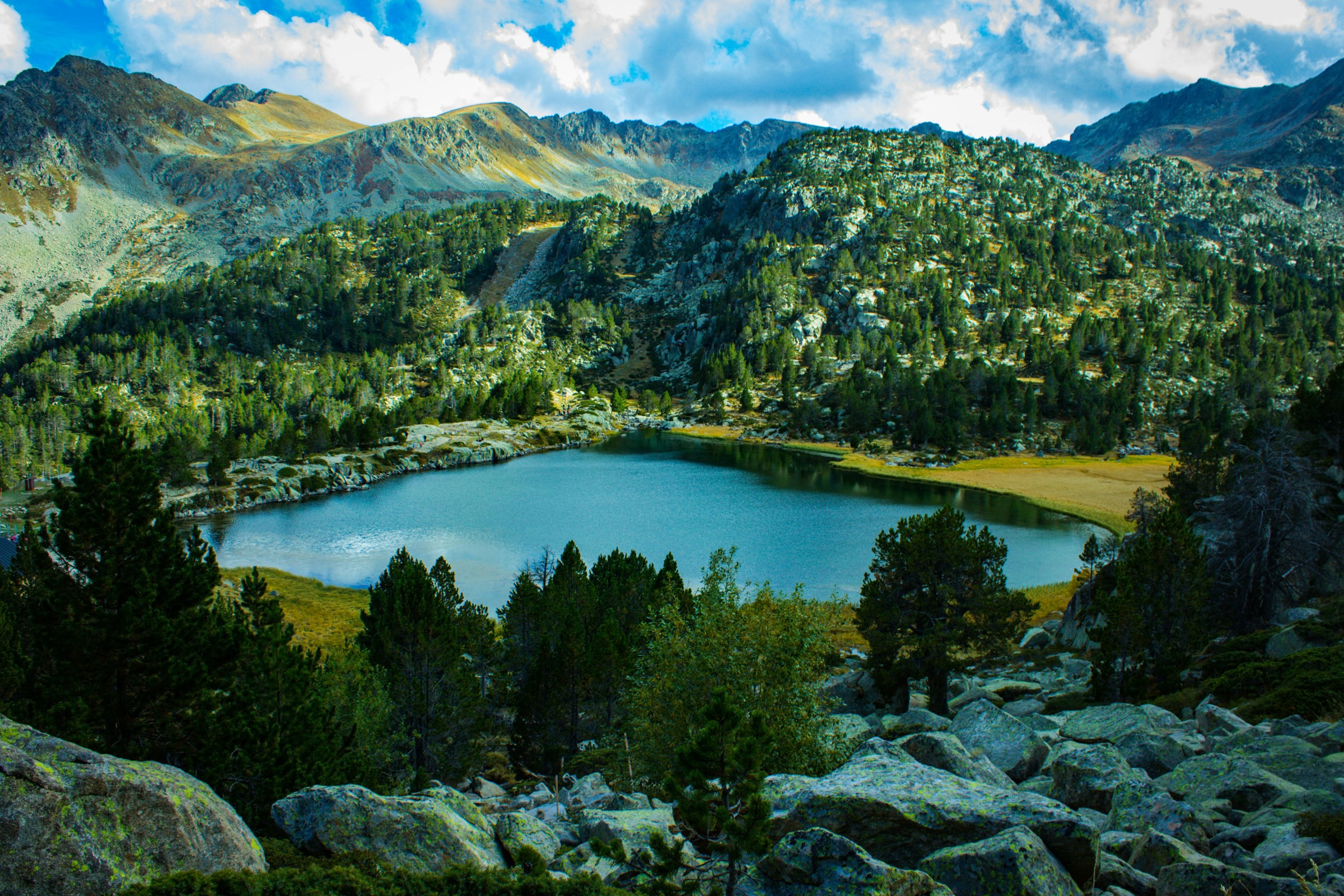 Bergsjö i Andorra omgiven av gröna träd och stenar med bergen i bakgrunden under en klar himmel.