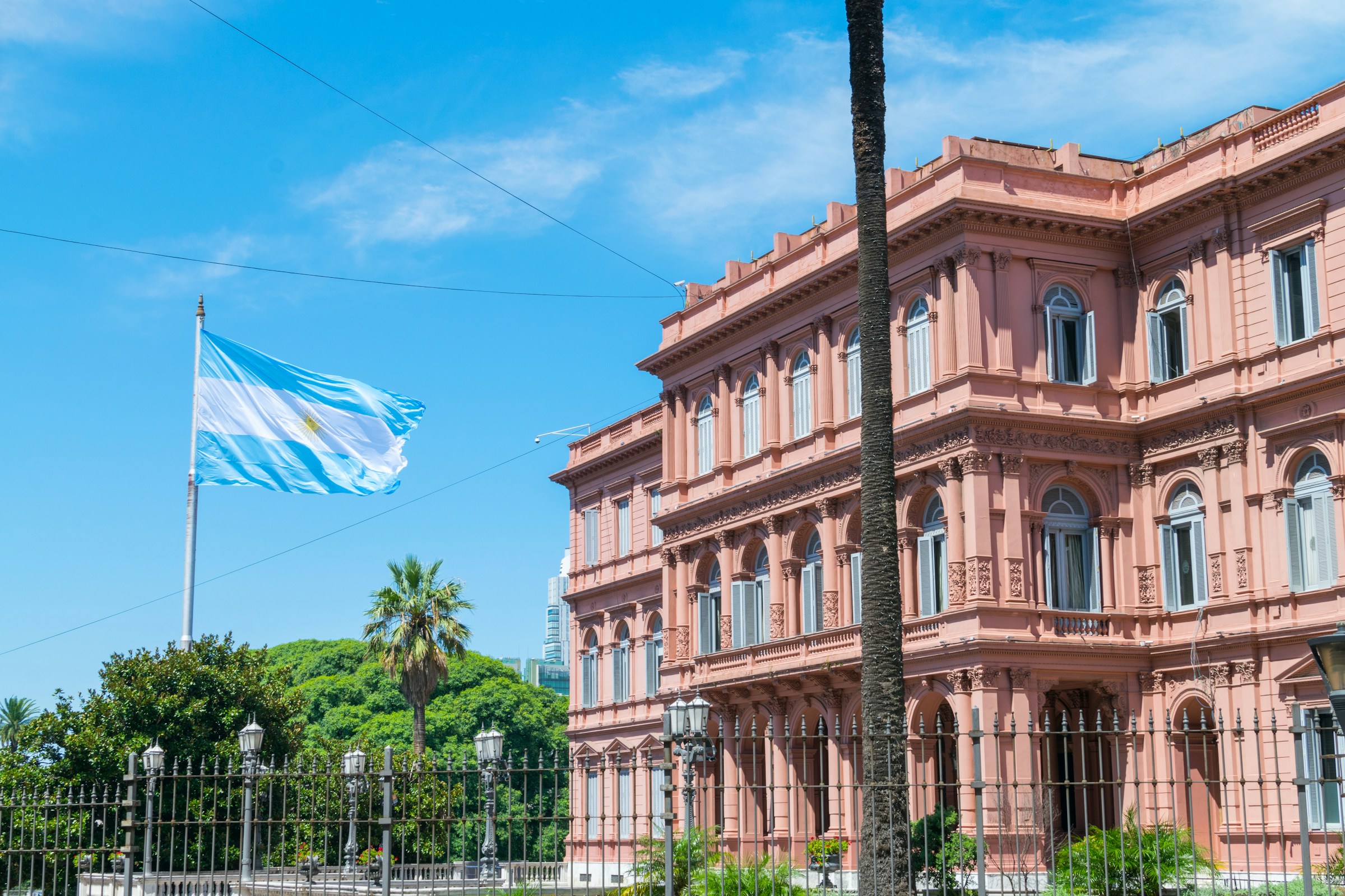 Argentinas flagga vajar framför presidentpalatset Casa Rosada i Buenos Aires under en klarblå himmel med grönska i förgrunden.