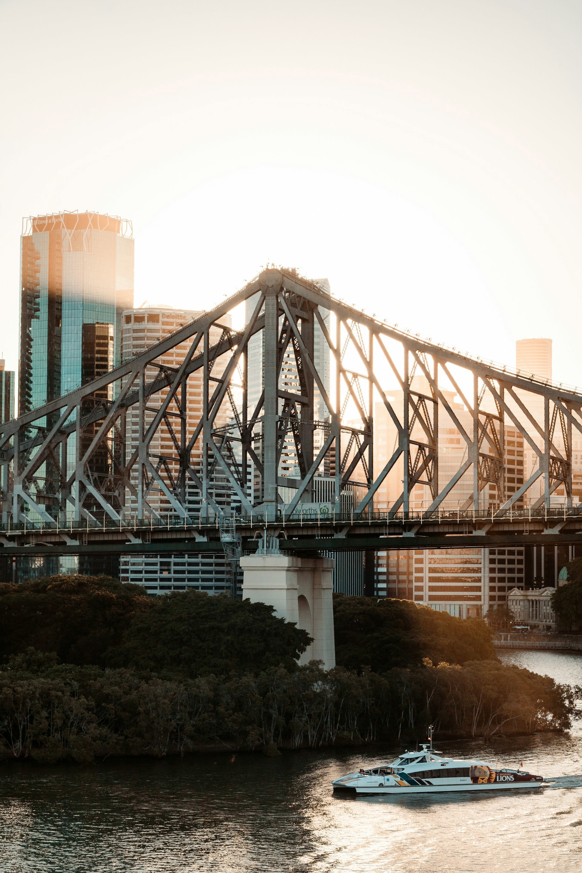 Solen skiner på en stålbro vid solnedgången med en färja som passerar under, omgiven av höghus och grönska i Brisbane.