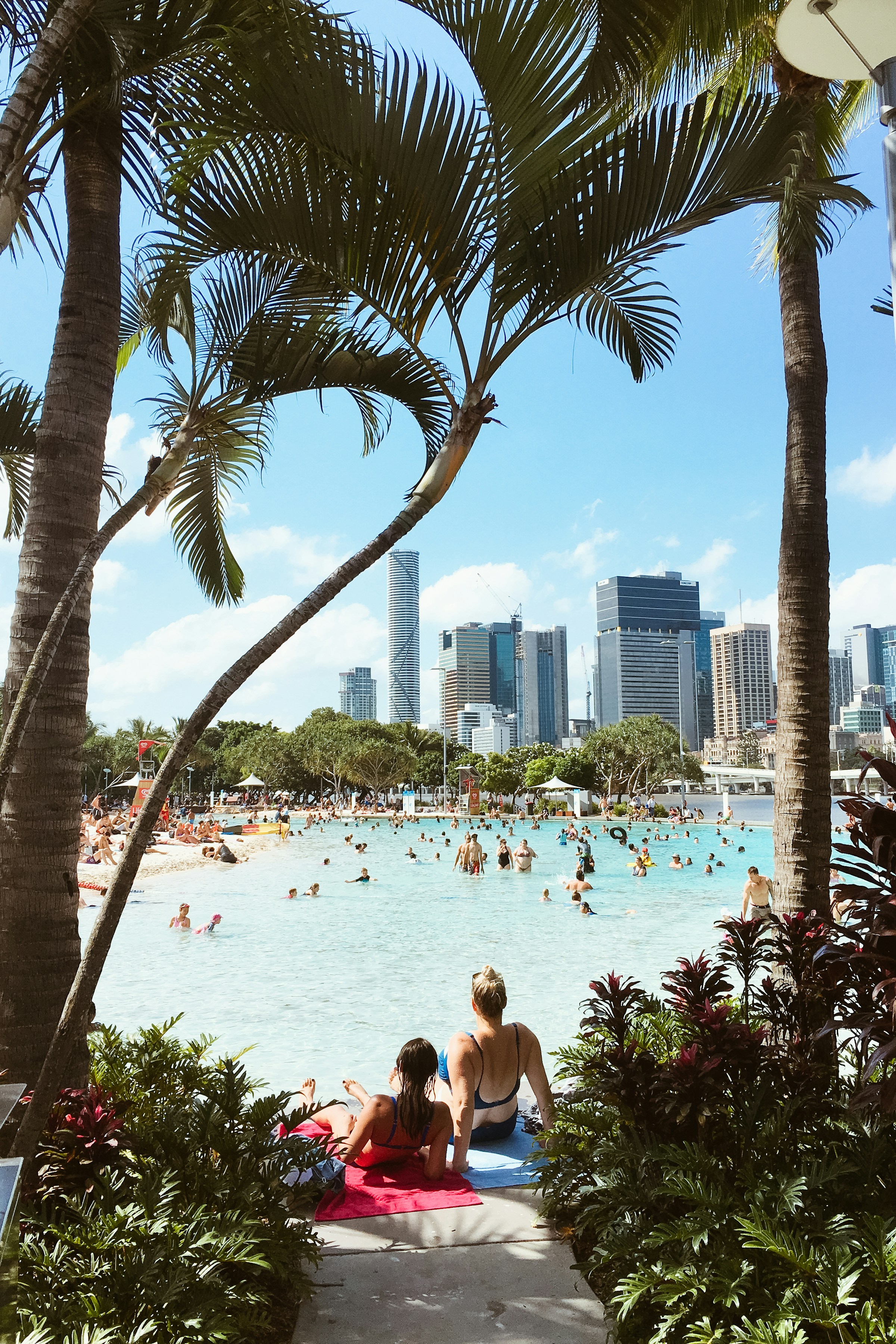 Stadsstrand med människor som badar och solar, omgiven av palmer, med stadssilhuett i bakgrunden på en solig dag I Brisbane.