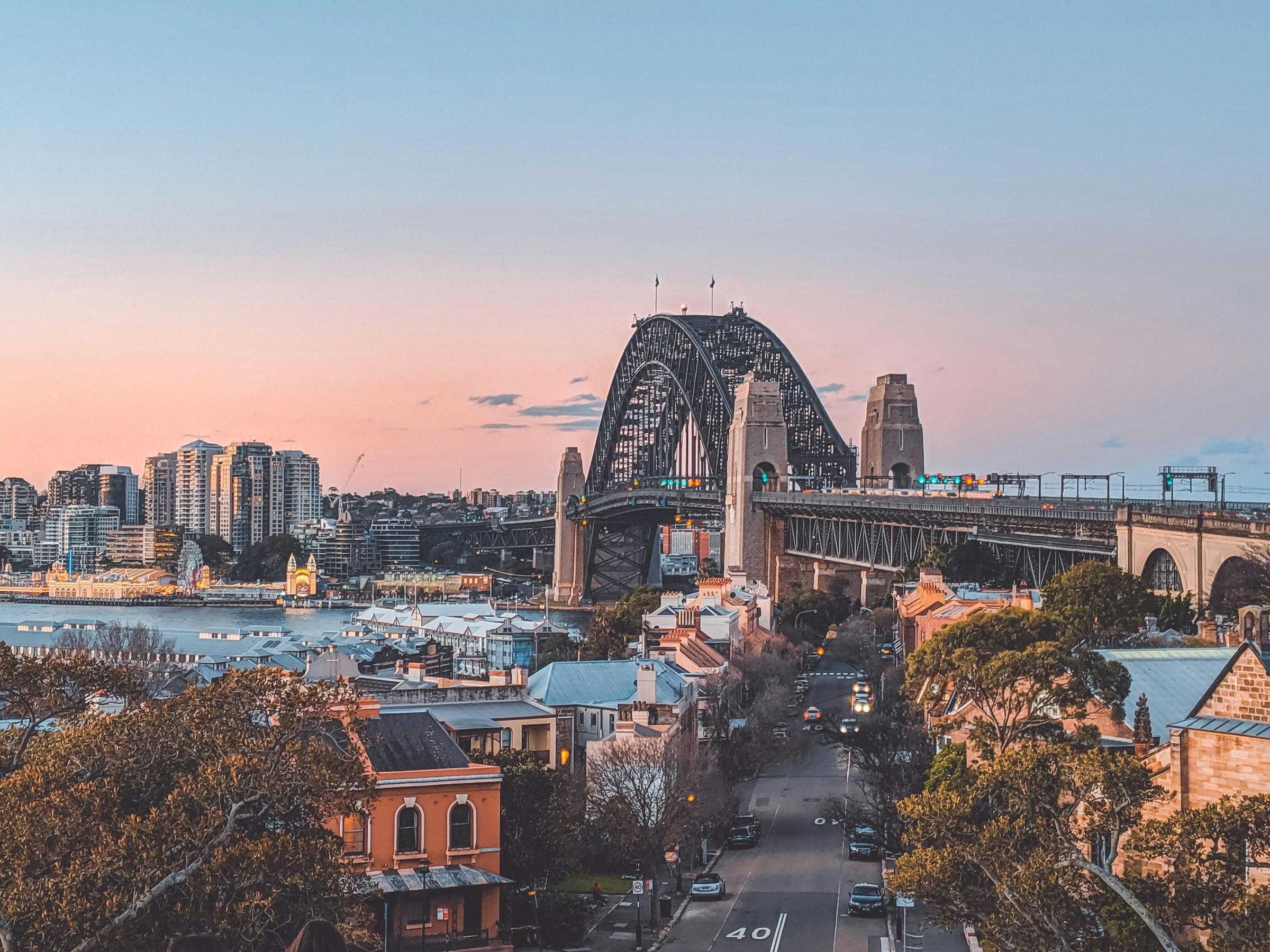 Vy över Sydney Harbour Bridge vid solnedgång med pastellfärgad himmel och stadssilhuett.