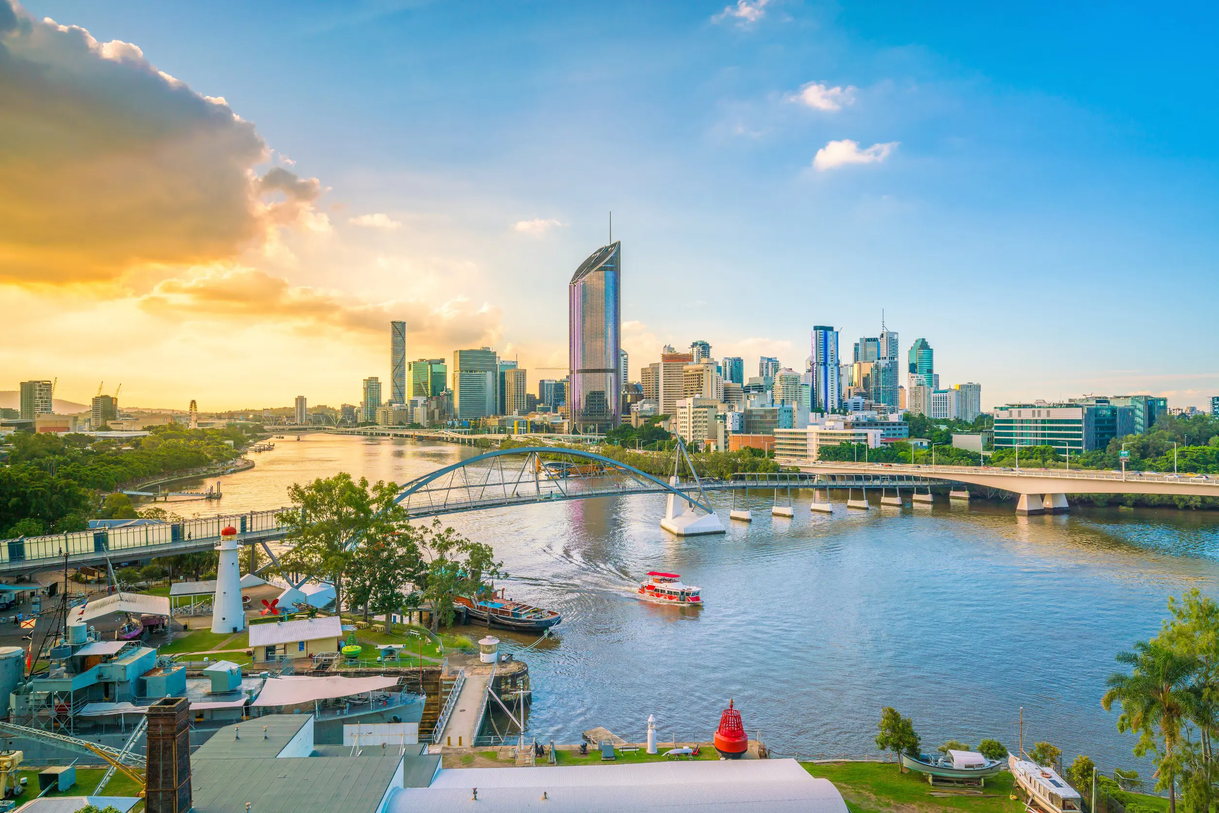 Resa till Brisbane - Vy över Brisbane stadskyline och Brisbane River vid solnedgång med blå himmel och lätta moln, synlig gångbro och färgglada byggnader.