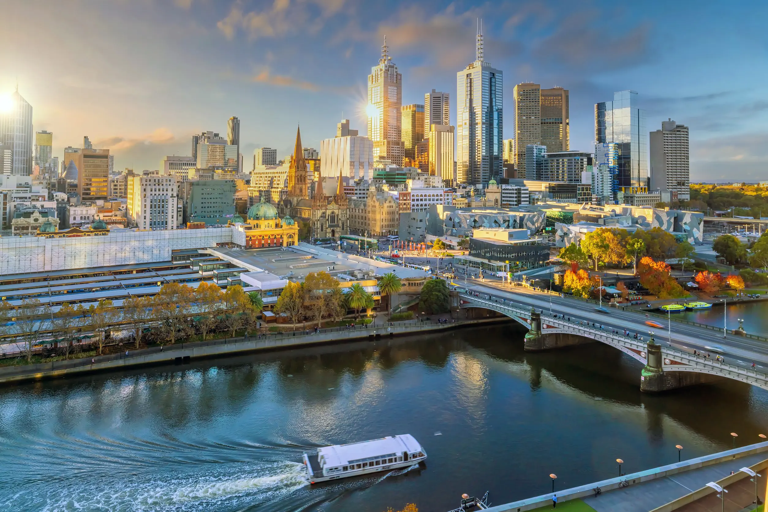 Resa till Melbourne - Solnedgång över Melbourne skyline med Yarra-floden och en flodbåt, en bro och frodiga träd vid vattnet.