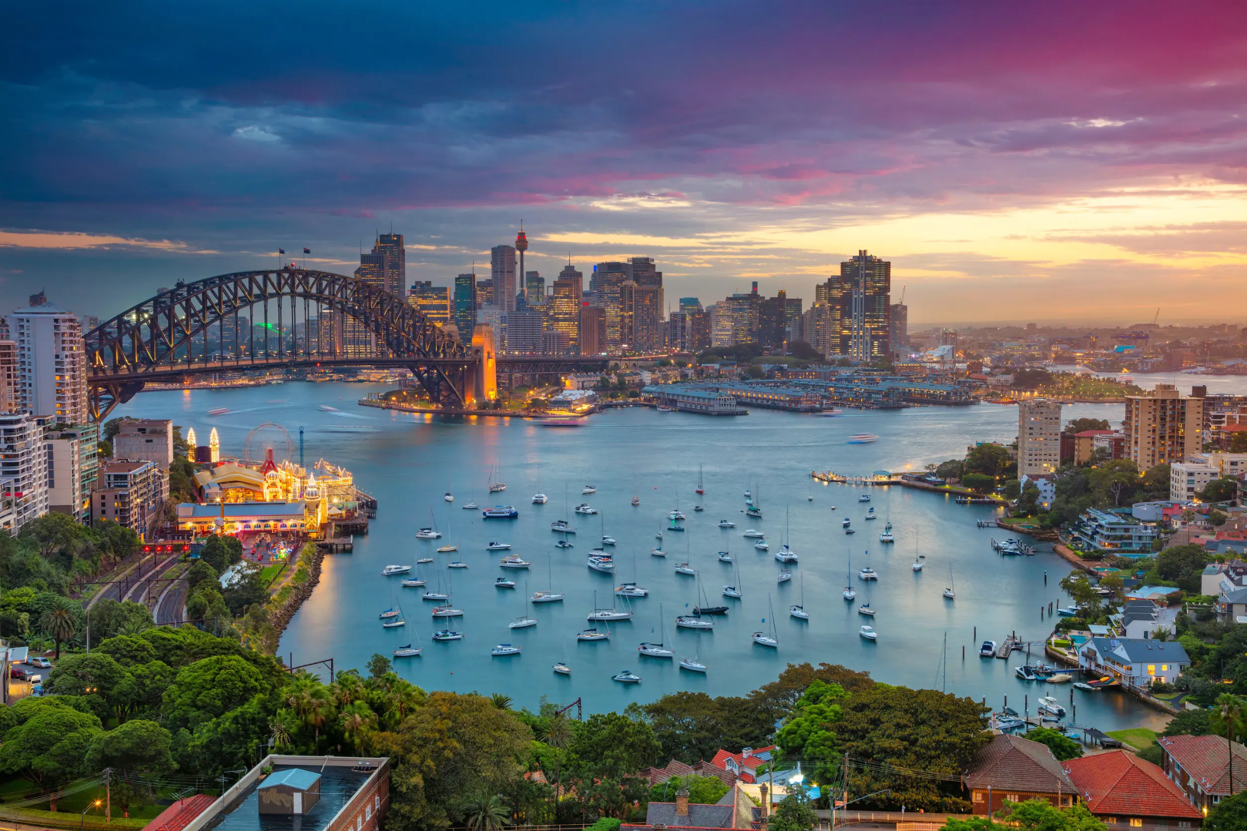 Resa till Sydney - Sydney hamn med Sydney Harbour Bridge vid solnedgång, stadssilhuett och båtar på vattnet.