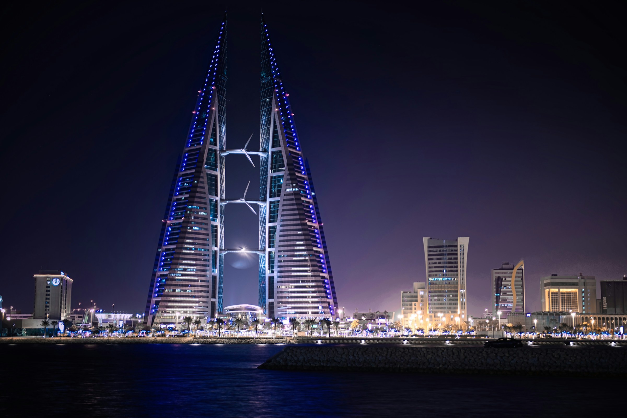 Upplyst skyskrapa med futuristisk design vid vattnet under nattetid med stadssilhuett i bakgrunden I Bahrain.