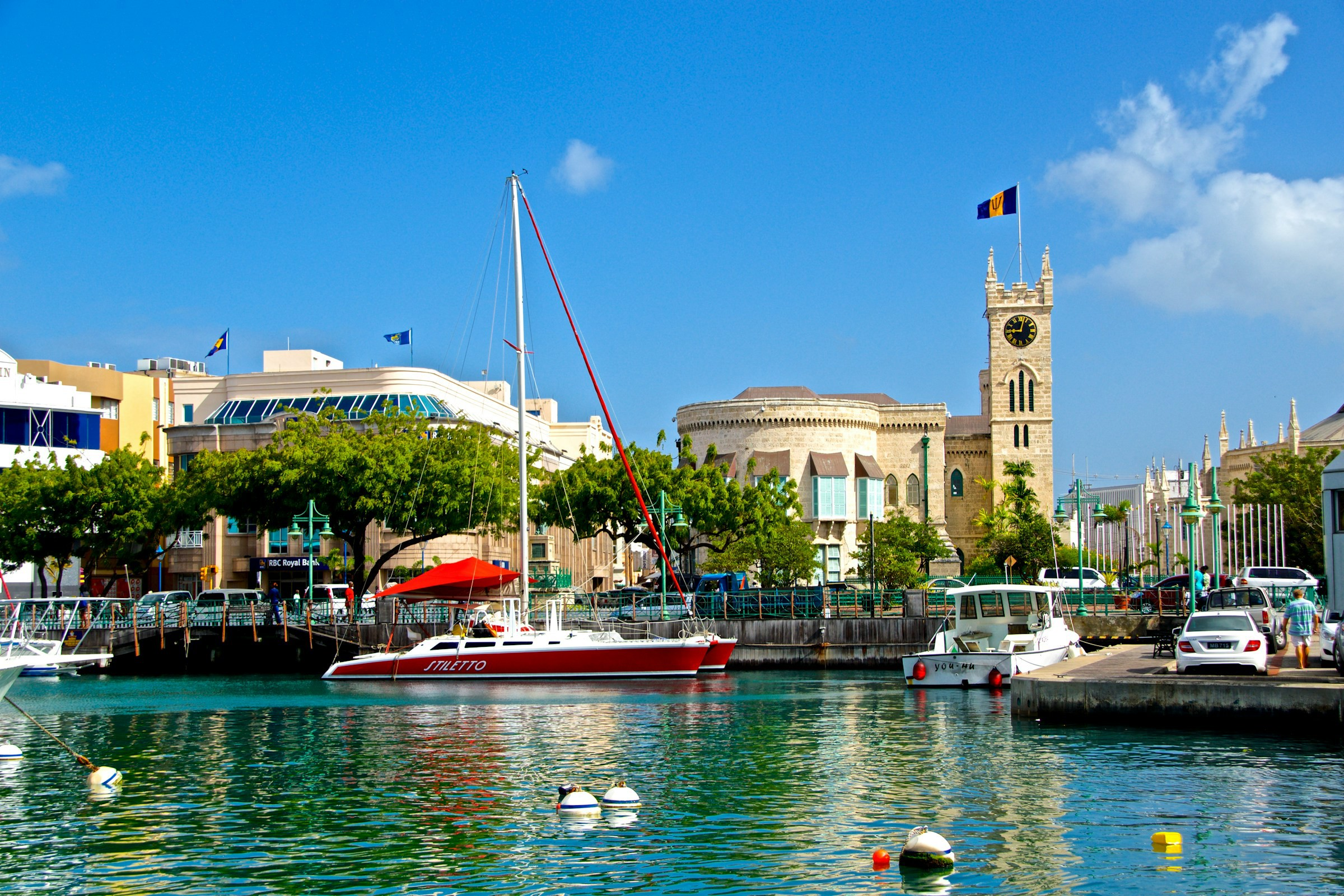 Livlig marina med färgglada båtar och reflekterande vatten framför historiska byggnader och ett klocktorn, en blå himmel med lätta moln på Barbados.