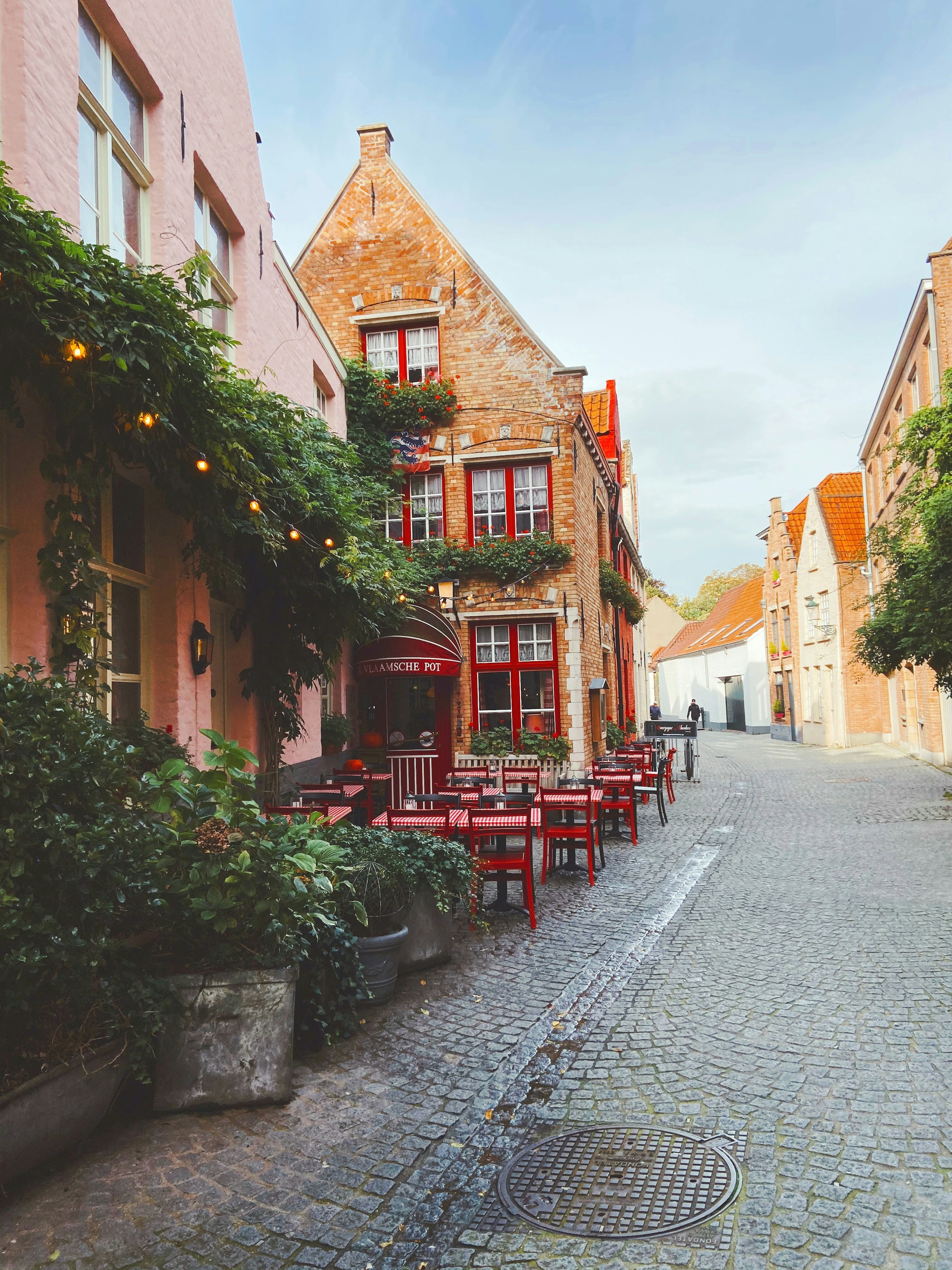 Mysig kullerstensgata med traditionell Belgisk arkitektur och uteservering med röda stolar och bord framför en historisk byggnad i Belgien.