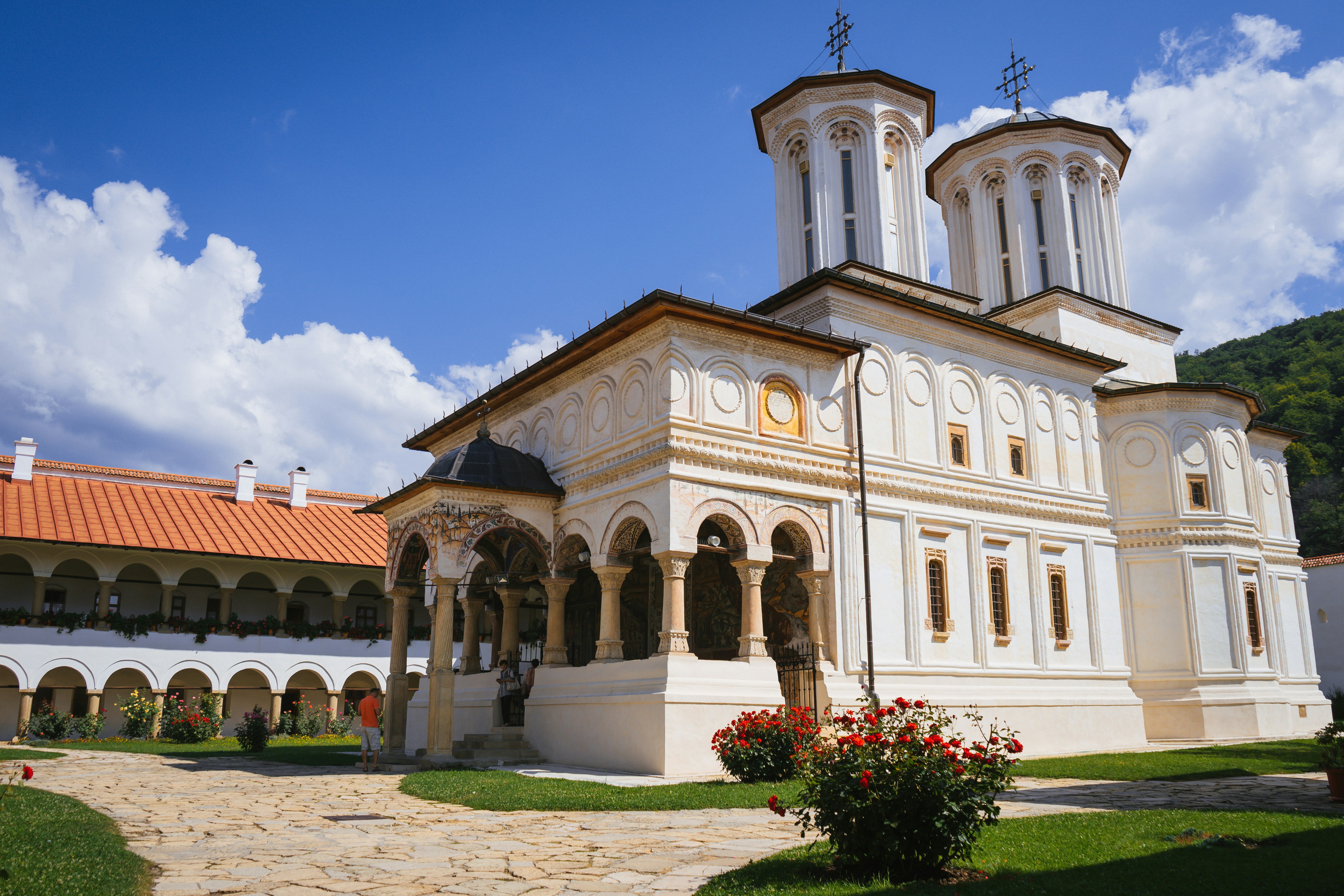 Vit klosterbyggnad med höga torn och arkadgångar, omgiven av lummig grönska och en klarblå himmel i Sarajevo.