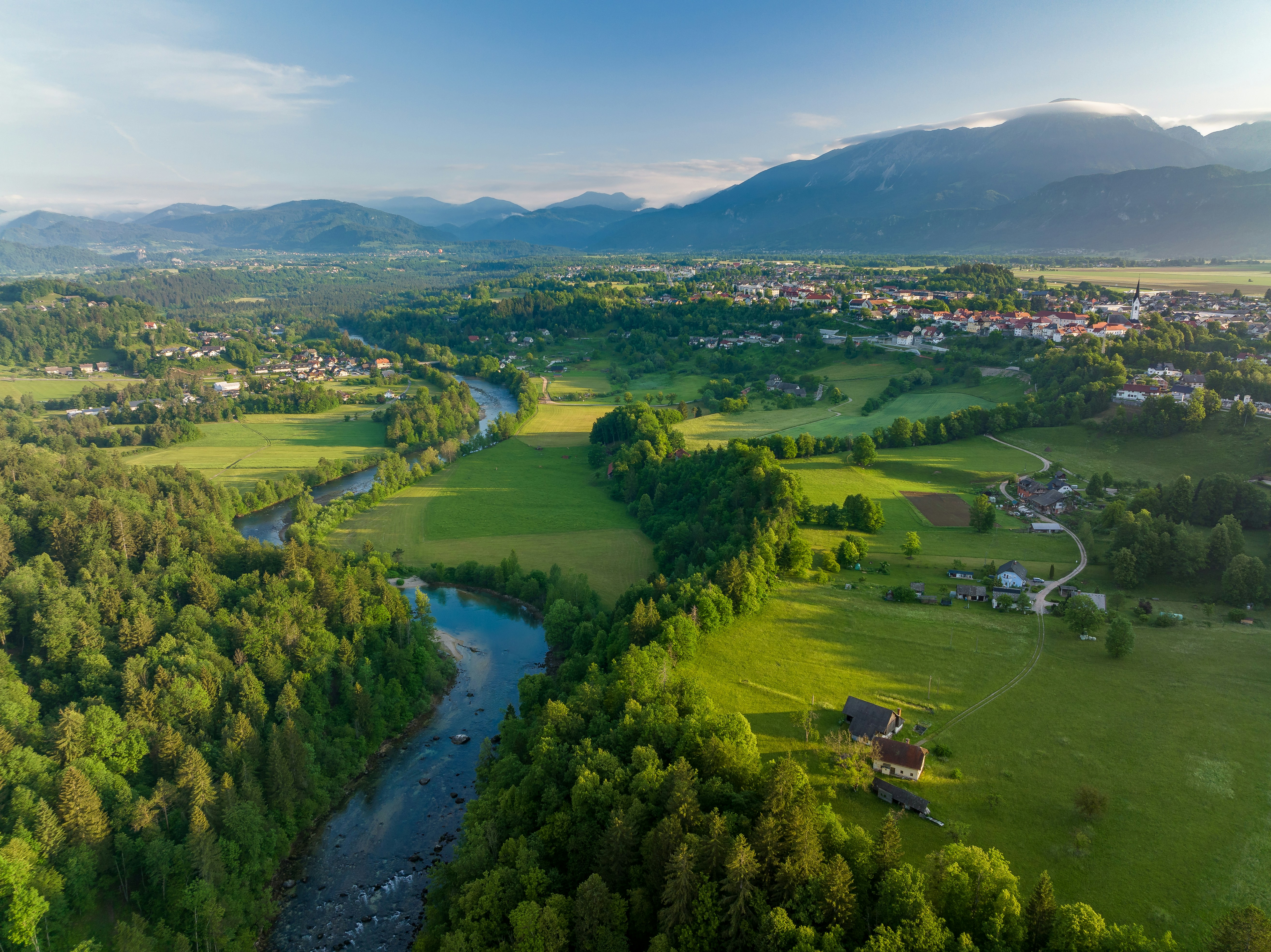 Flygfoto över lantlig idyll med slingrande flod, gröna fält och by i bakgrunden med berg i Bosnien.
