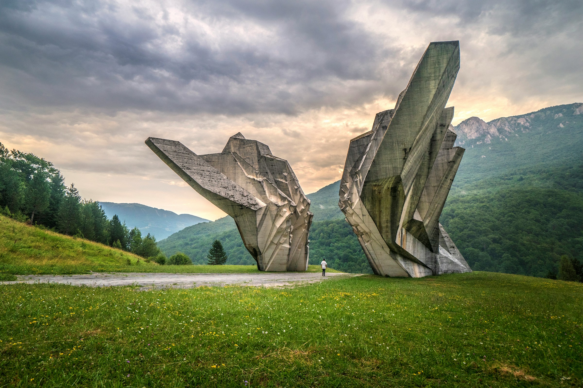 Monumental skulptur i betong på grönt fält med kullar i bakgrunden och en person som betraktar konstverket i Bosnien.