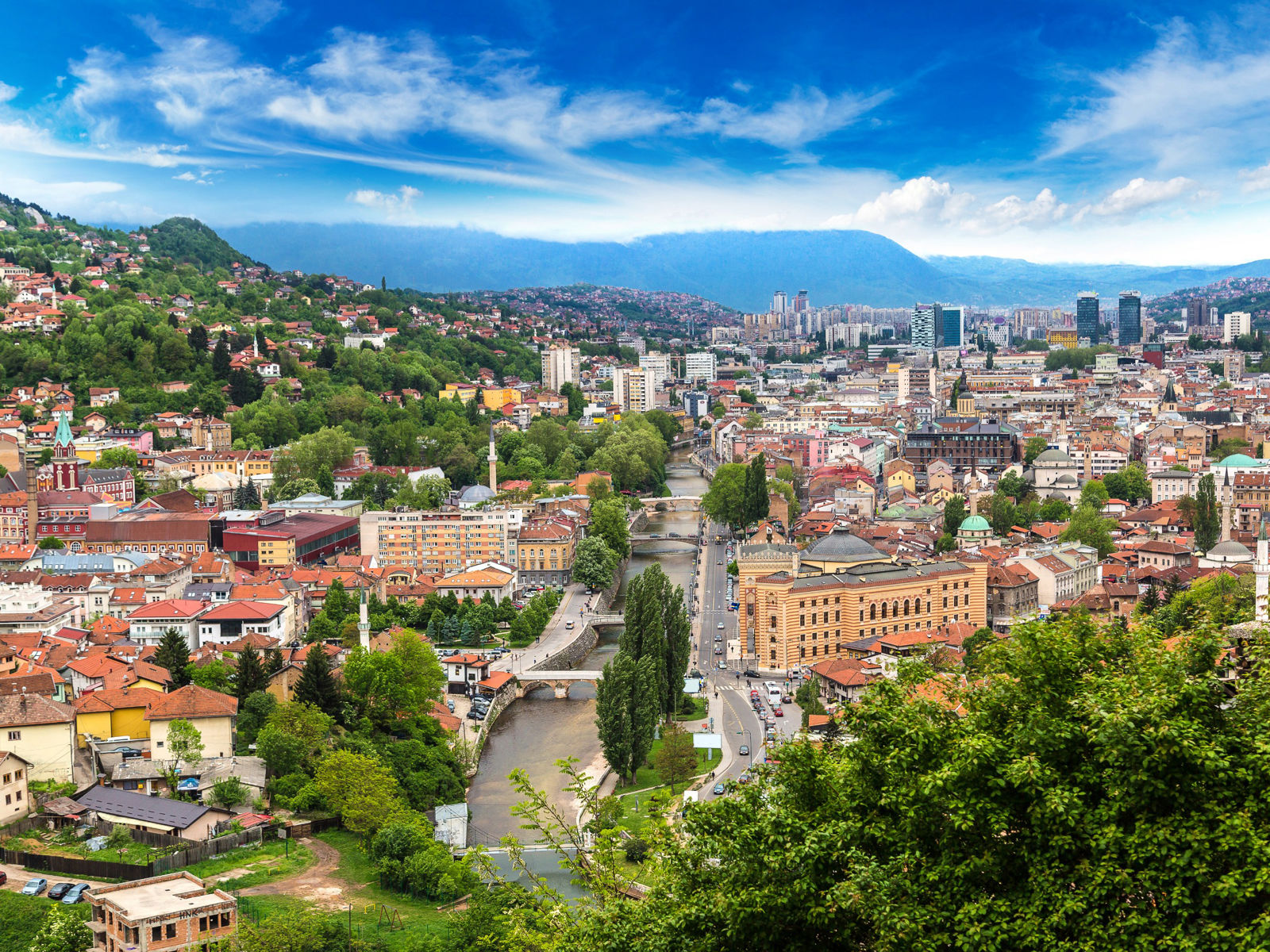 Resa till Sarajevo - Panoramautsikt över en livlig stad med byggnader, flod och gröna kullar under en molnig himmel.