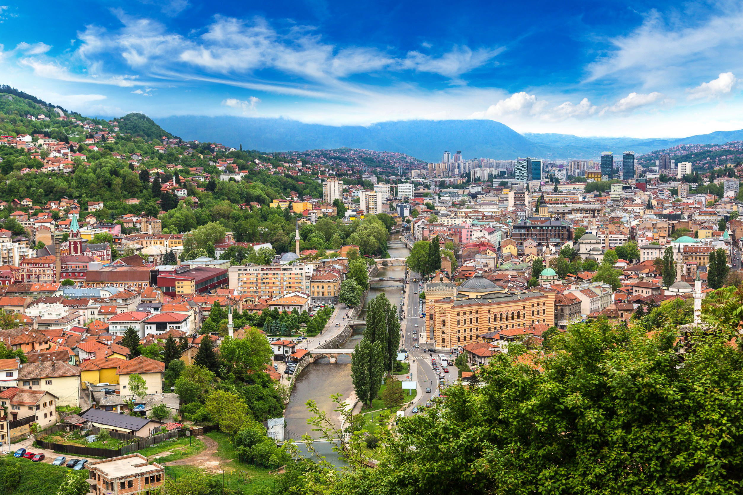 Resa till Sarajevo - Panoramautsikt över en livlig stad med byggnader, flod och gröna kullar under en molnig himmel.