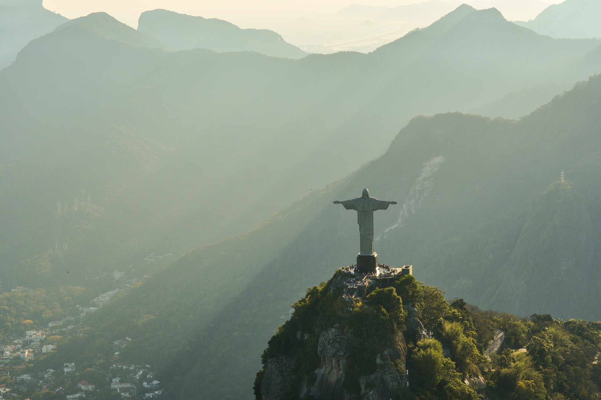 Vy över berömd Kristusstaty på bergstoppi Rio De Janeiro under solnedgång med lummiga kullar i bakgrunden.