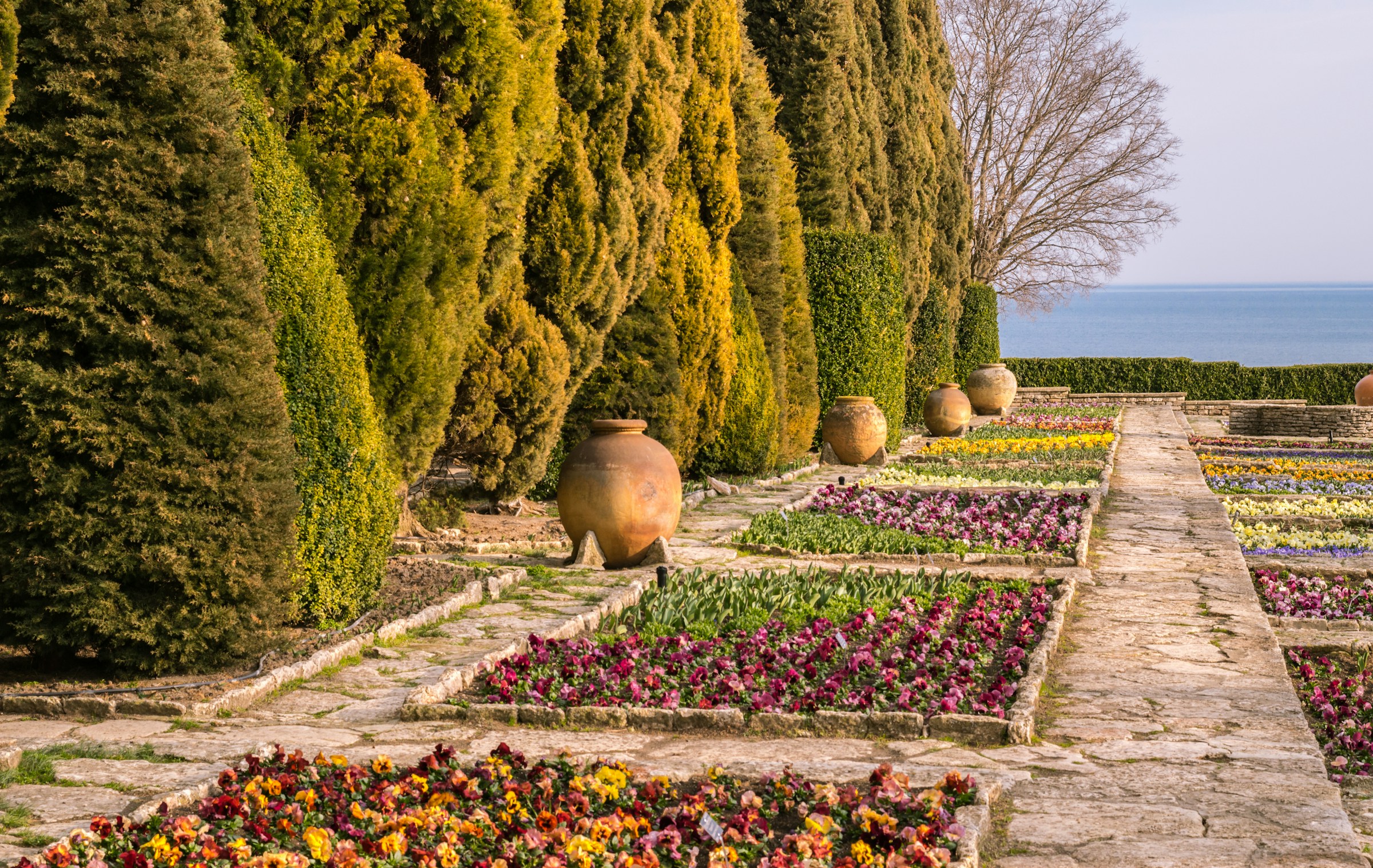 En välskött trädgård med mönstrade blomsterrabatter i olika färger längs en stenlagd gångväg, traditionella lerkrukor och en bakgrund av hav och himmel i Varna.