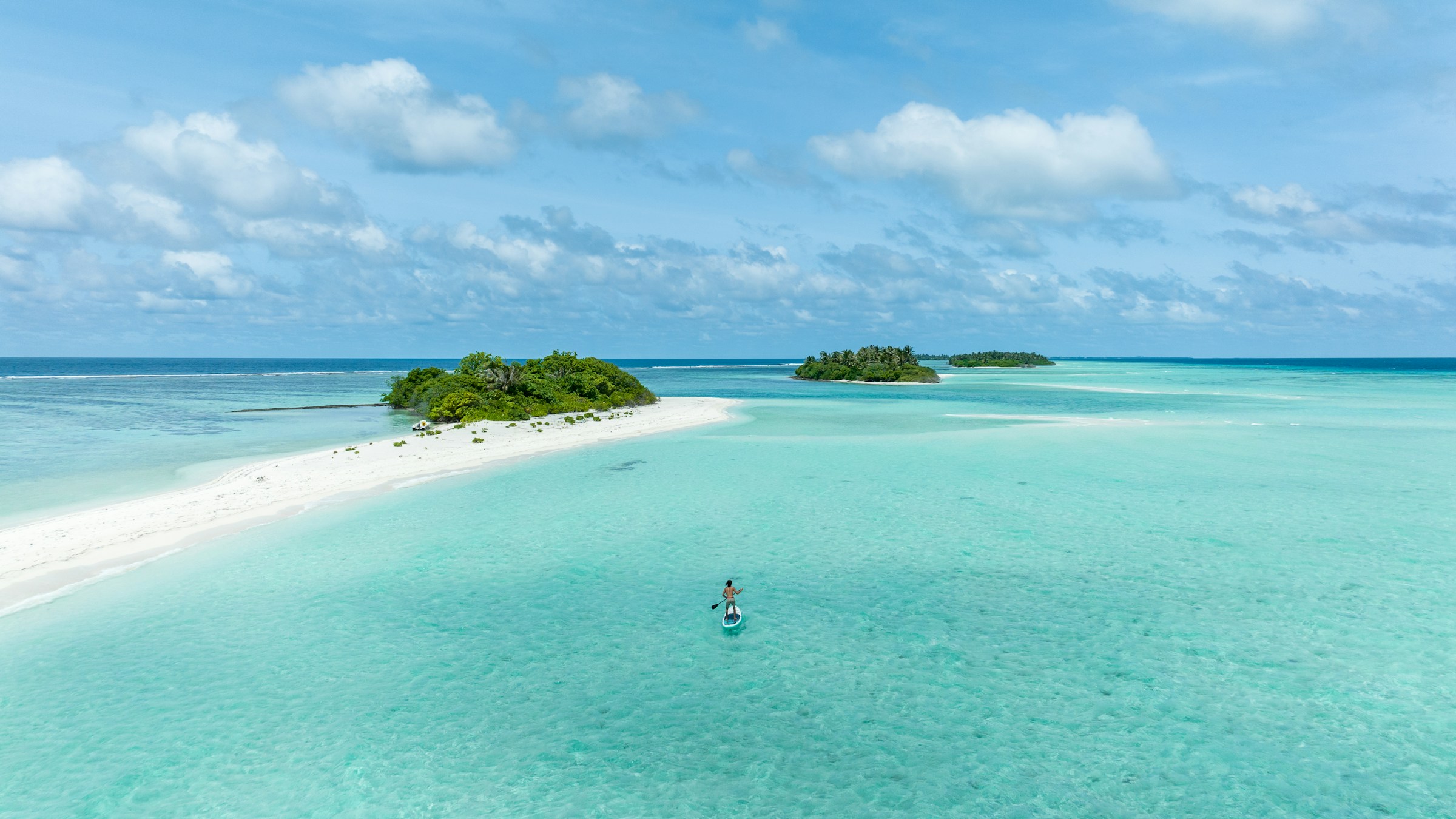 Vy över tropiska öar med kristallklart turkos vatten och en person som paddlar på en standuppaddlebräda, vita sandstränder och frodig grönska i bakgrunden på Caymanöarna.