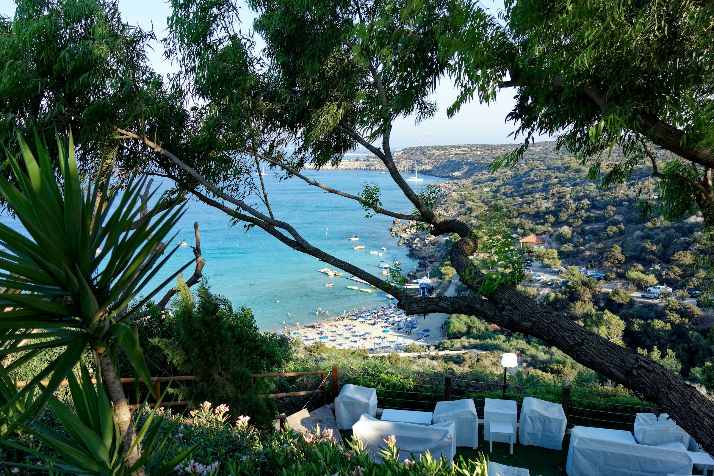 Utsikt över en idyllisk vik med klart blått vatten och båtar från en lummig trädgård med medelhavsgrönska och vita utemöbler på en solig dag i Ayia Napa.