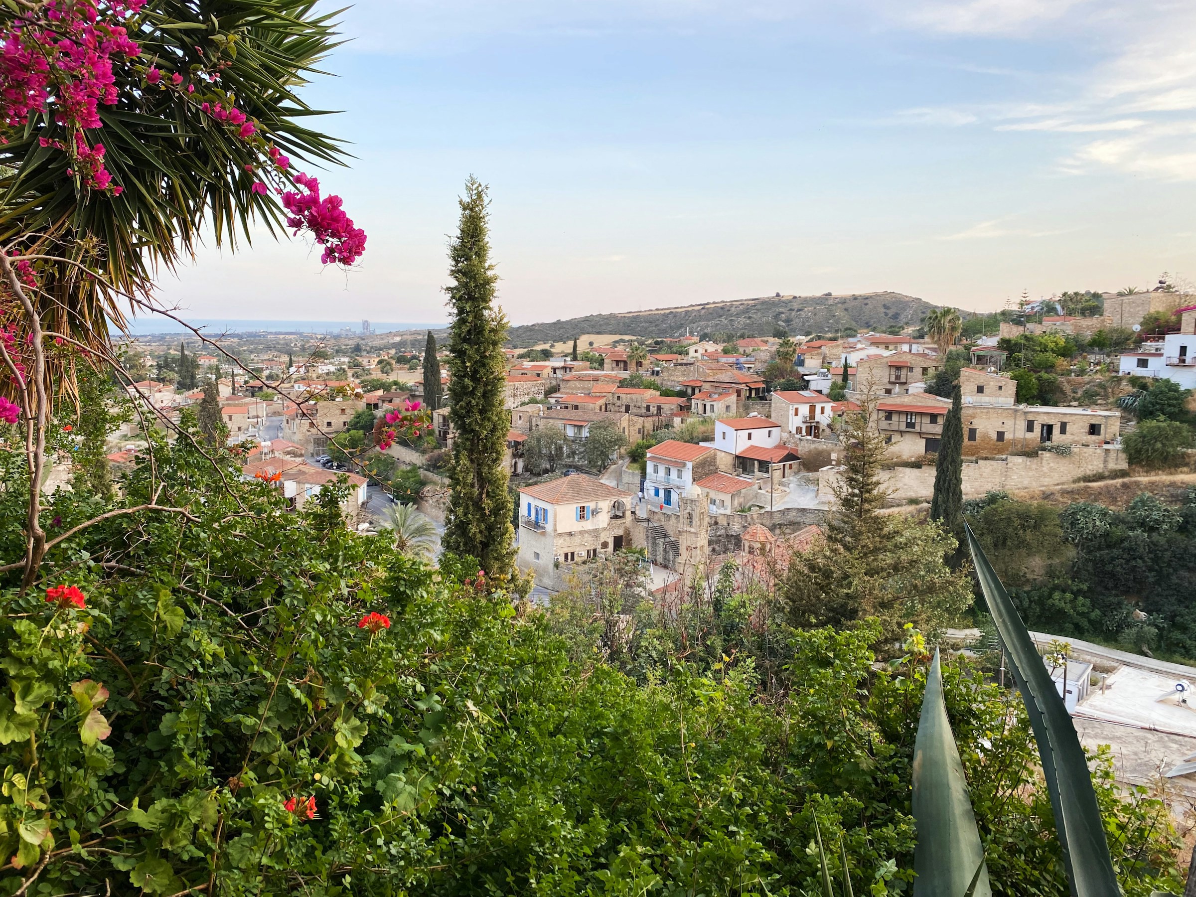 Utsikt över en pittoresk by med traditionella hus, gröna träd och blommande buskar i förgrunden, med kullar i bakgrunden vid skymning i Larnaca.