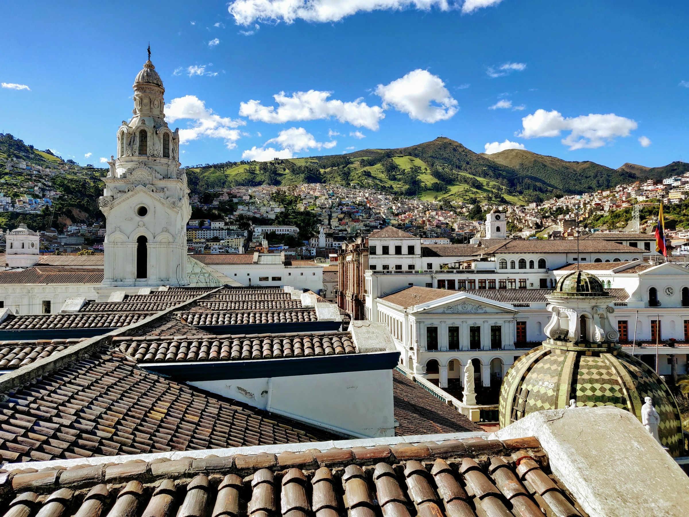 Vy över historisk stadskärna i Ecuador med kolonial arkitektur och takpannor i förgrunden, en vit kyrktorn mot en klarblå himmel och kuperade gröna kullar i bakgrunden.