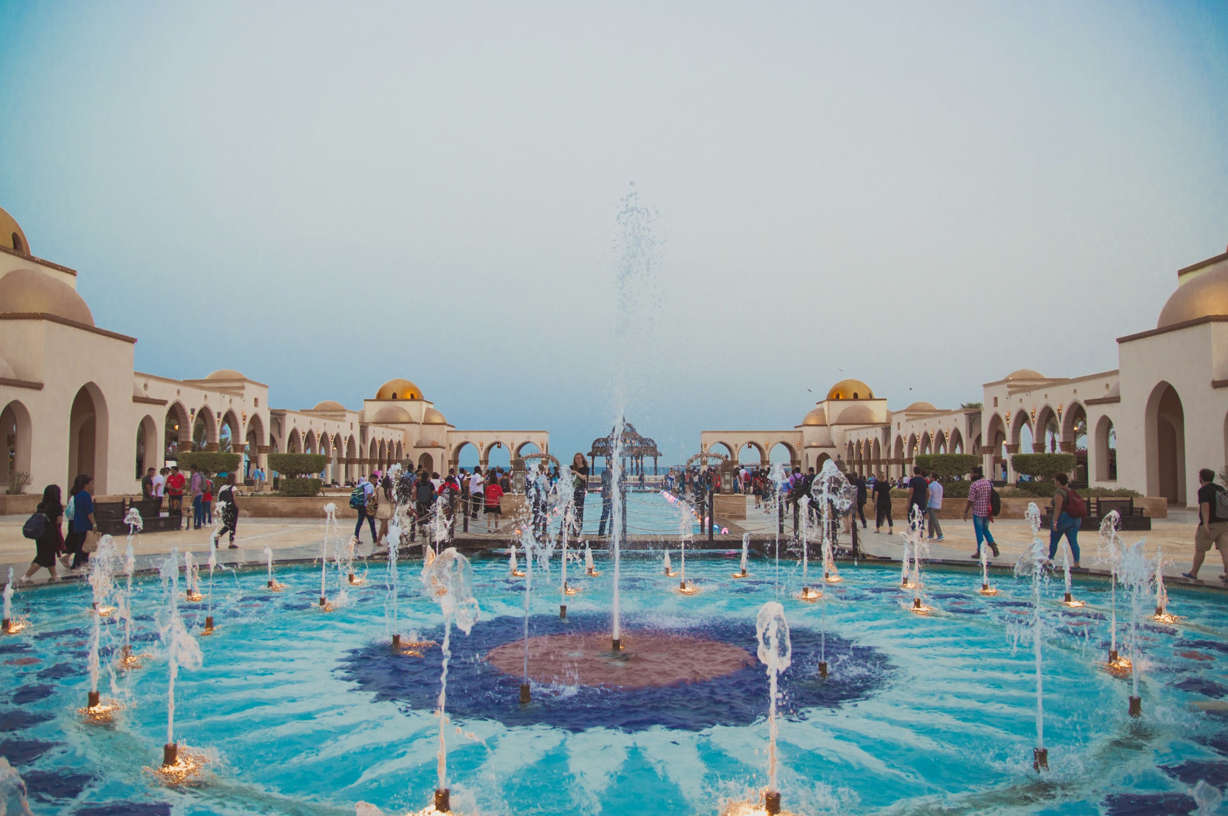 Folk promenerar kring en dekorativ fontän i en arkadgång med kupolbyggnader vid skymning i Hurghada.