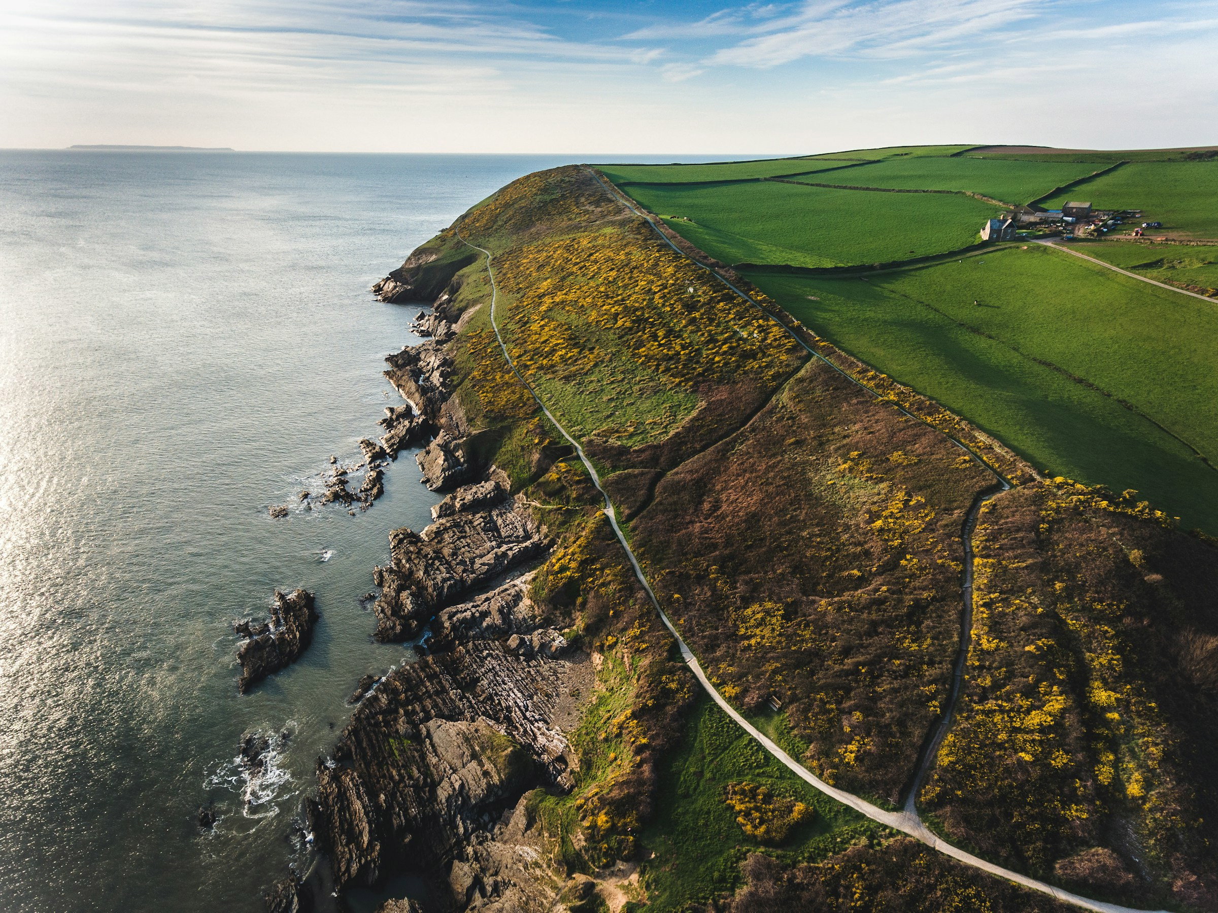 Luftbild av en kustnära klippig udde med gröna fält och gula blommor vid havet i England.