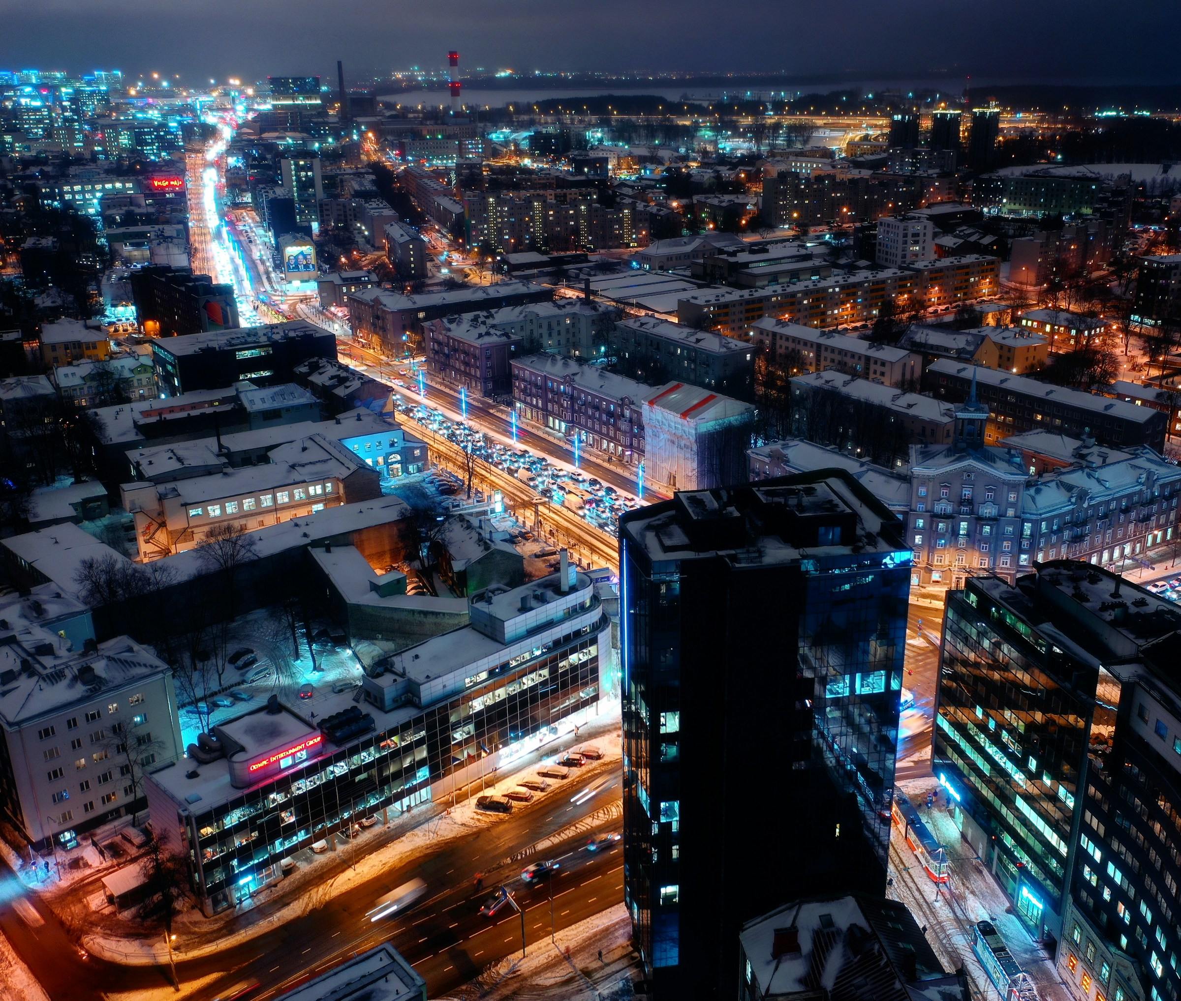 Flygfoto över en upplysta Tallinn på natten med gator som lyser i neon, snötäckta byggnader och aktiv trafik, som framhäver stadens liv och rörelse.
