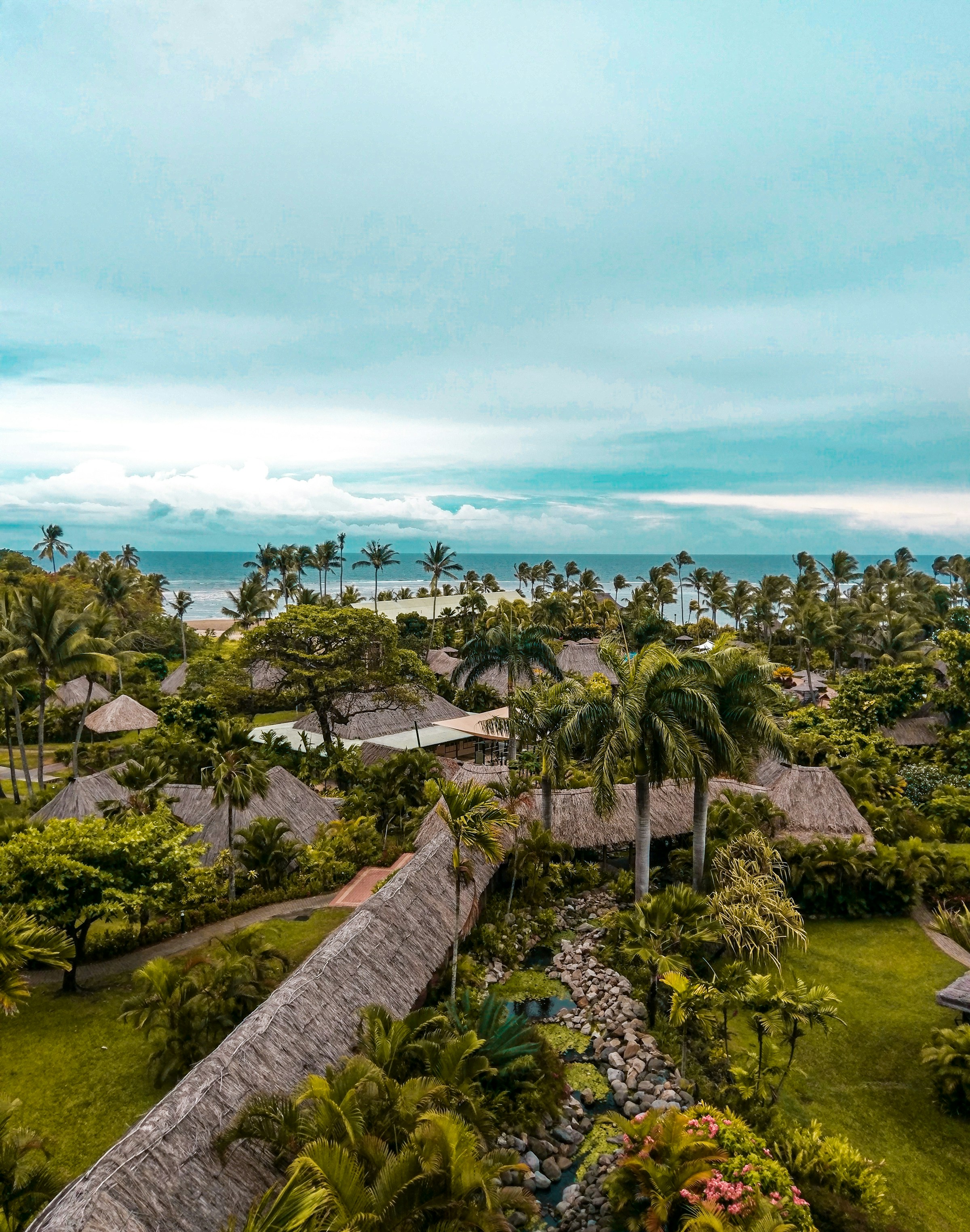 Tropisk resort med grästakade bungalows omgiven av grönska, palmträd och en ström, med havsutsikt och moln på himlen på Fiji.