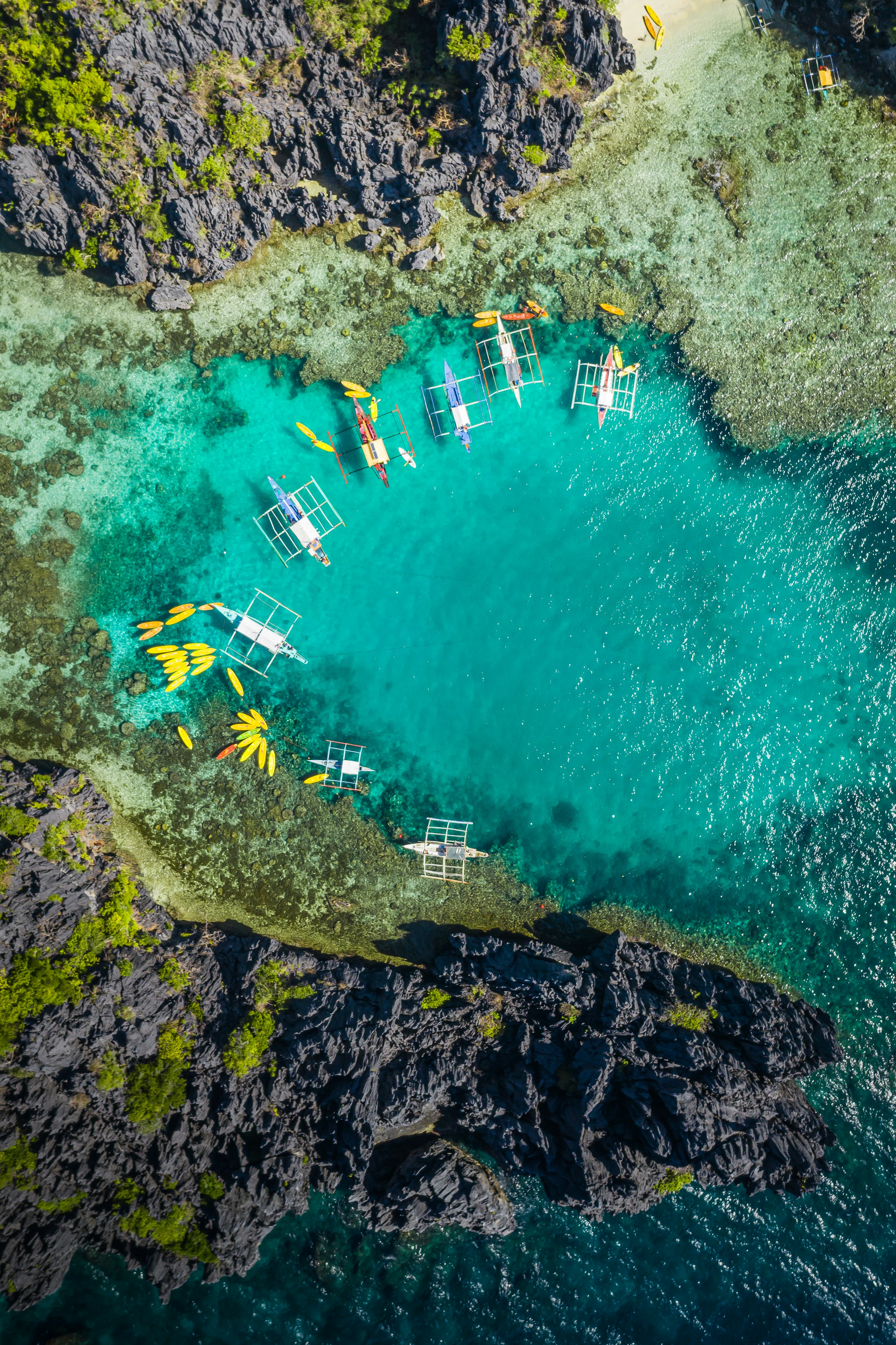 Flygbild av kristallklart turkost vatten med kajaker och traditionella båtar nära klippiga kusten på Filippinerna.