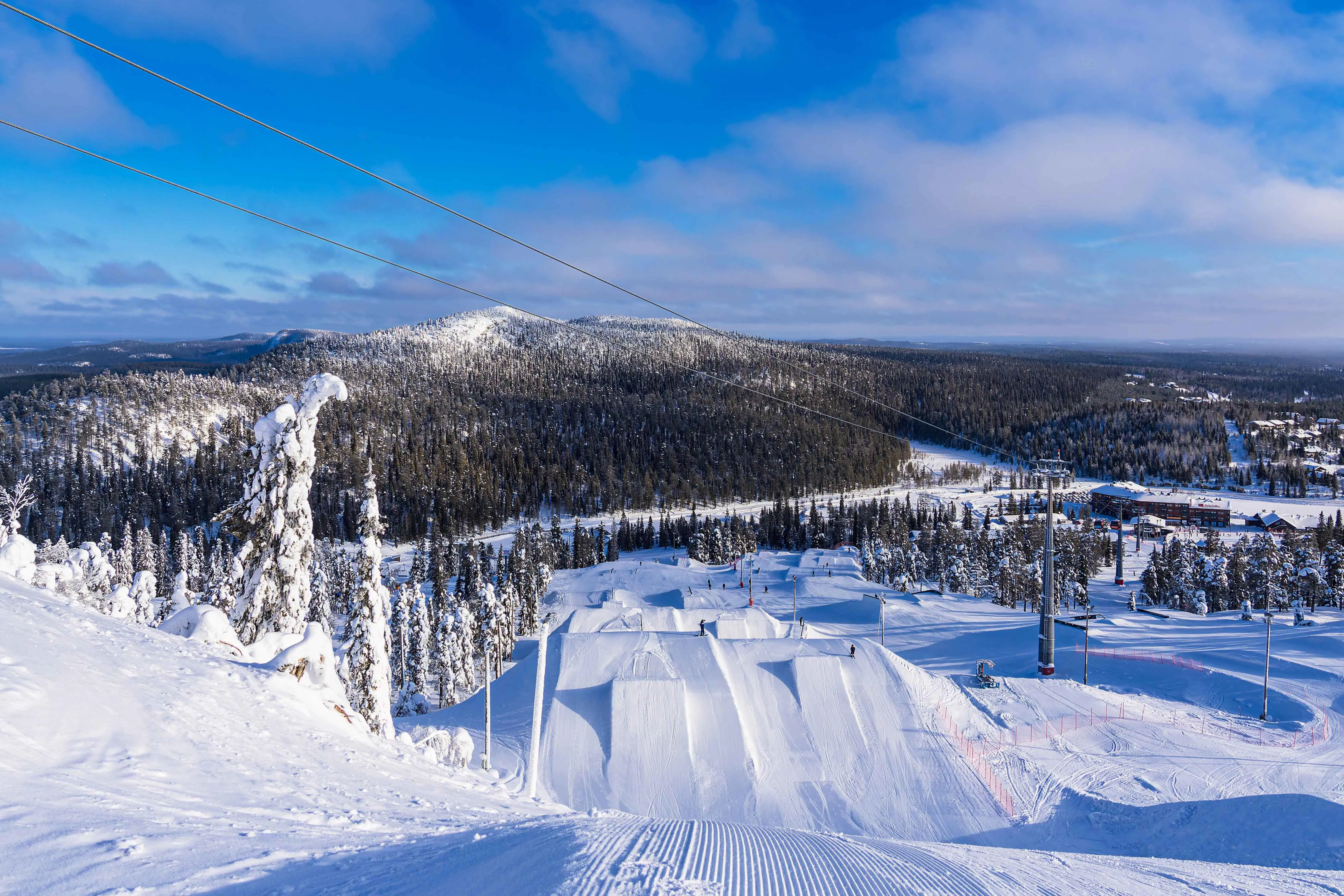 Resa till Ruka - Snötäckt skidbacke leder ner mot tät skog och snö med blå himmel i bakgrunden i Finland