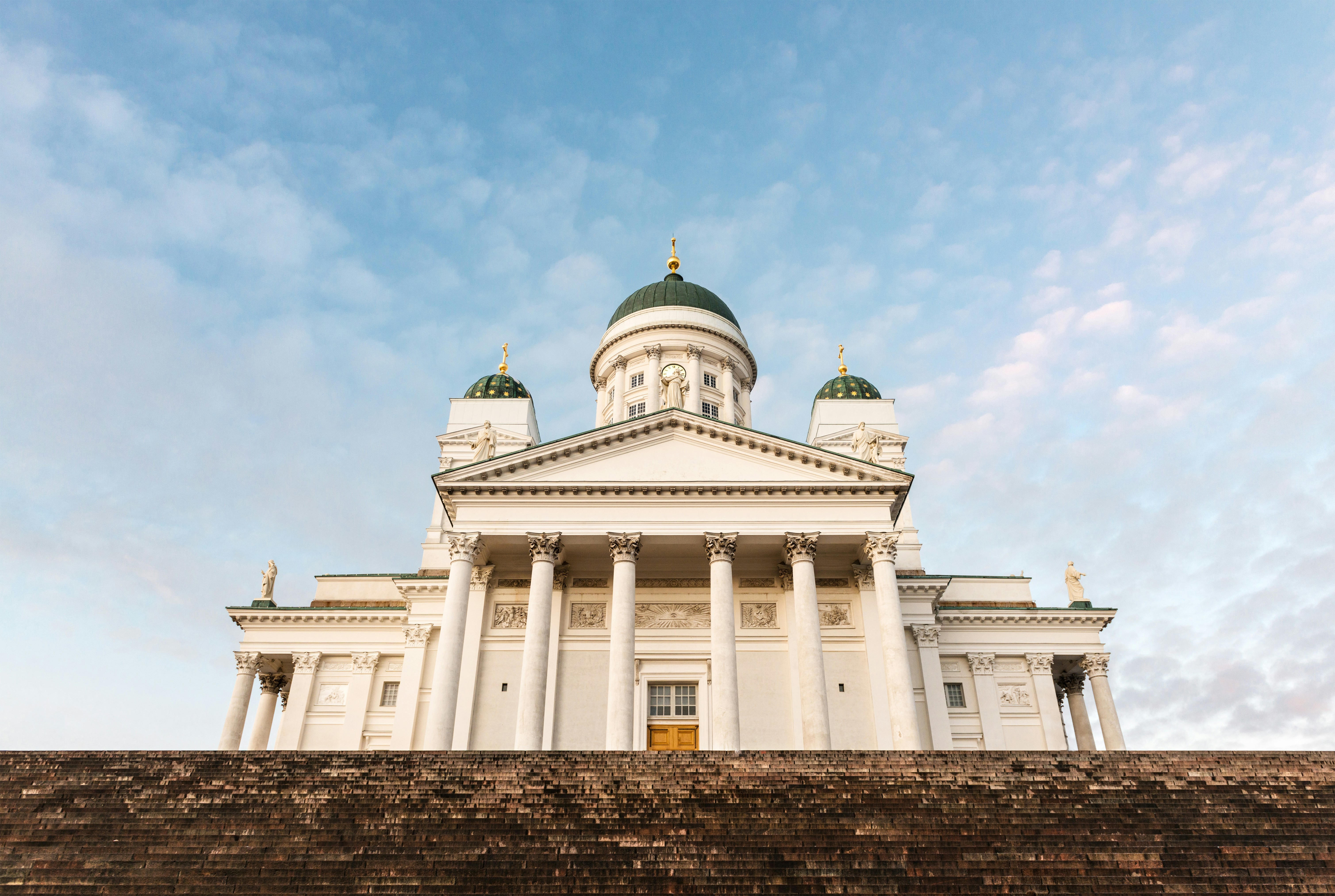 Vy över Helsingfors domkyrka i Finland vid solnedgång med dess imponerande vita fasad och gröna kupoler mot en klarblå himmel.